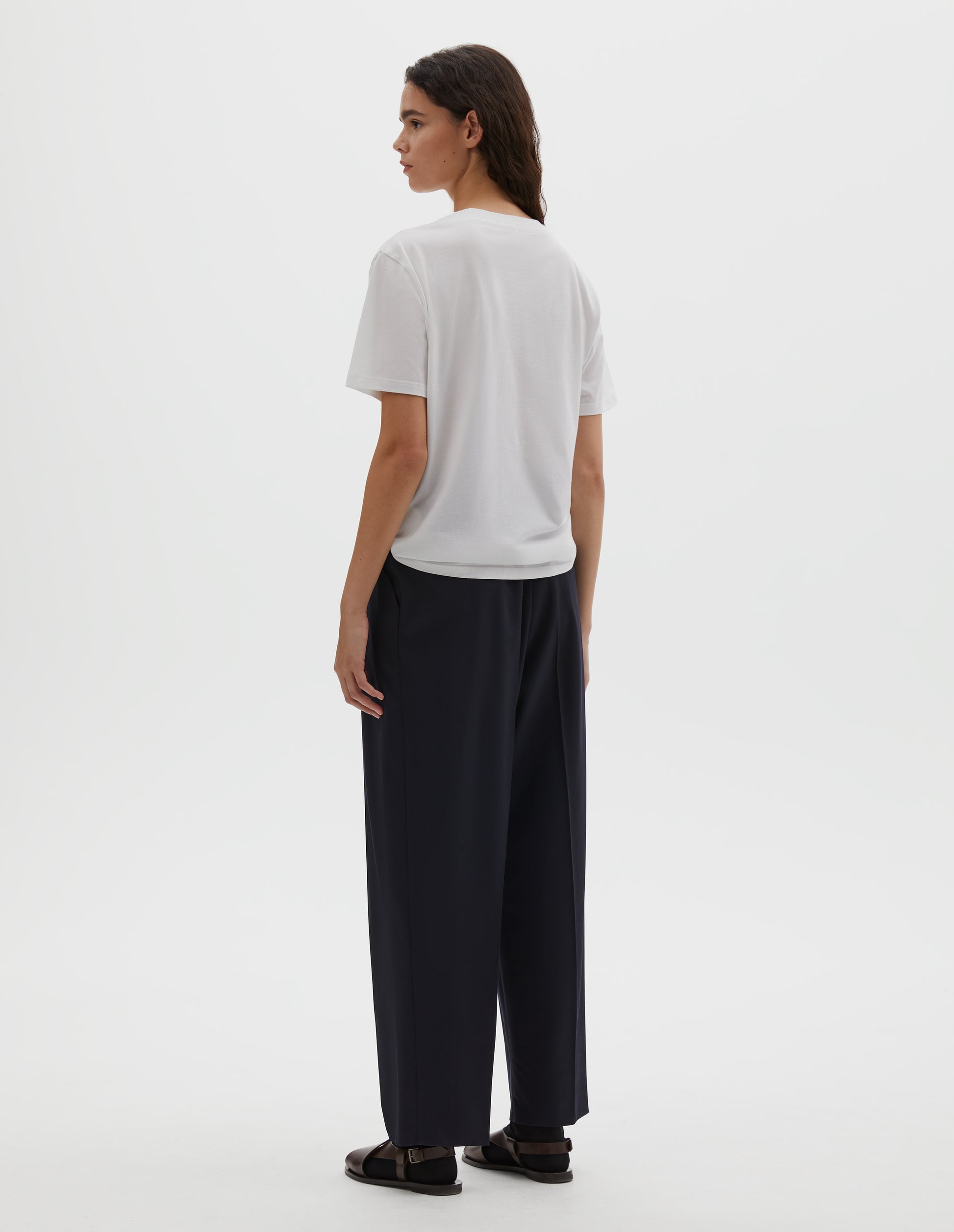 Full body image, back view of model wearing off white cotton short sleeve t-shirt with boxy cut, Styled with dark navy pleated trousers, black socks and brown leather sandals.