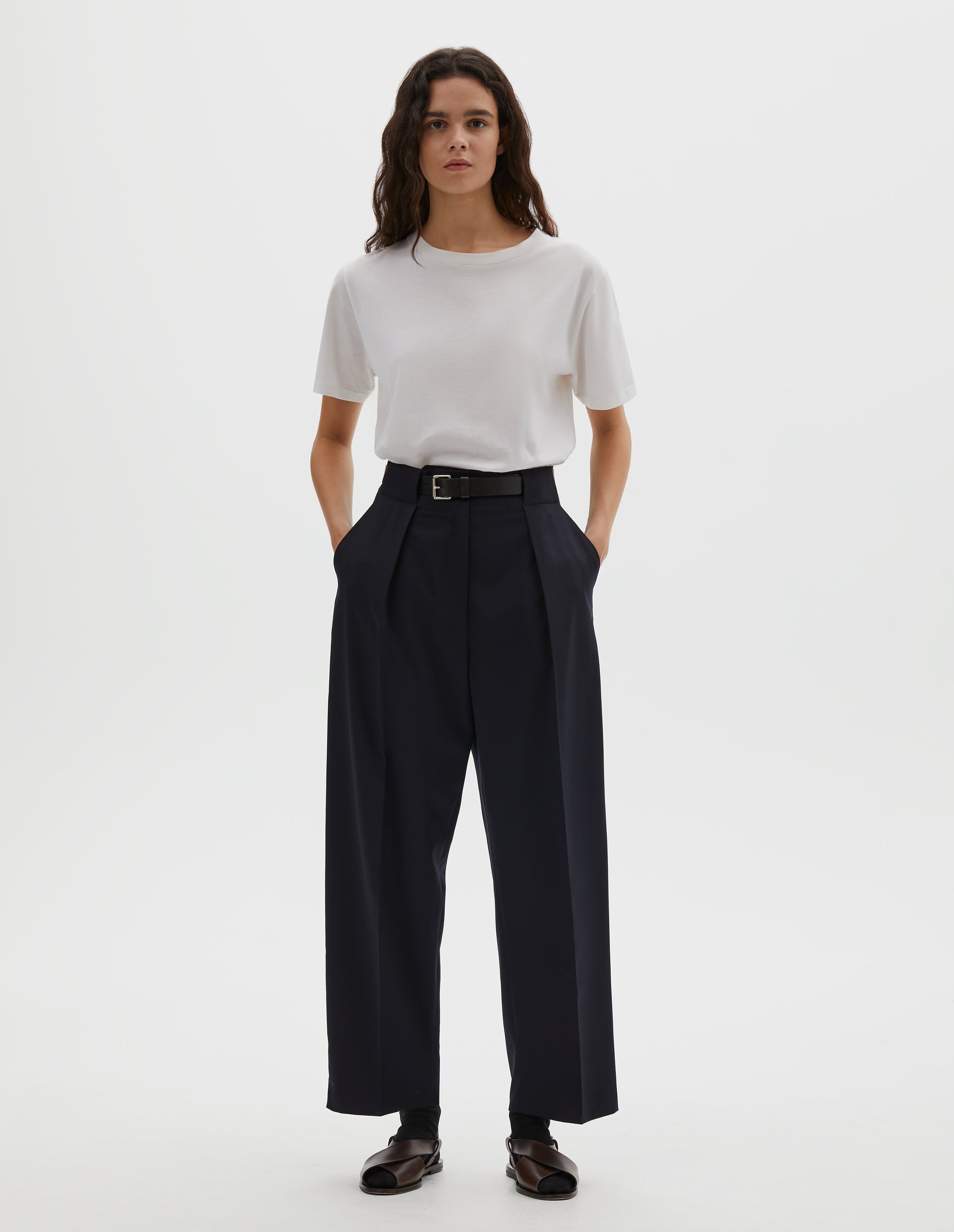 Full body image, front view of model wearing dark navy wool trousers, single pleats and grown-on waistband with large belt loops and horn button. Styled with white t-shirt, blue socks and brown leather sandals.