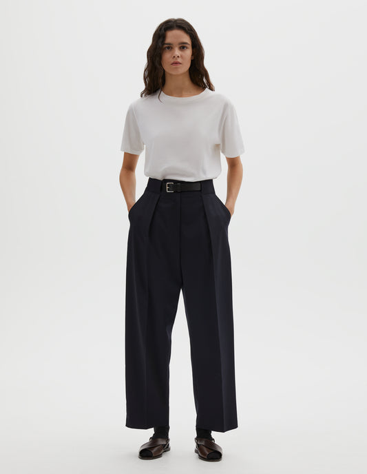 Full body image, front view of model wearing dark navy wool trousers, single pleats and grown-on waistband with large belt loops and horn button. Styled with white t-shirt, blue socks and brown leather sandals.
