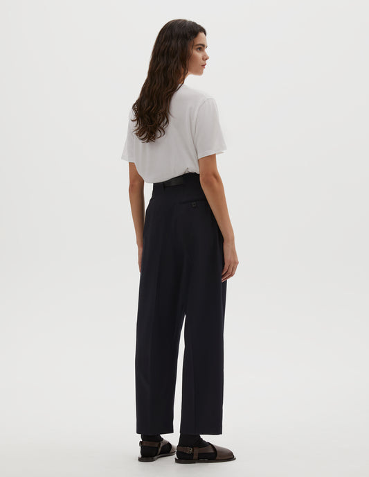 Full body image, back view of model wearing dark navy wool trousers, grown-on waistband with large belt loops and a button-through jet back pocket and horn button. Styled with white t-shirt, blue socks and brown leather sandals.