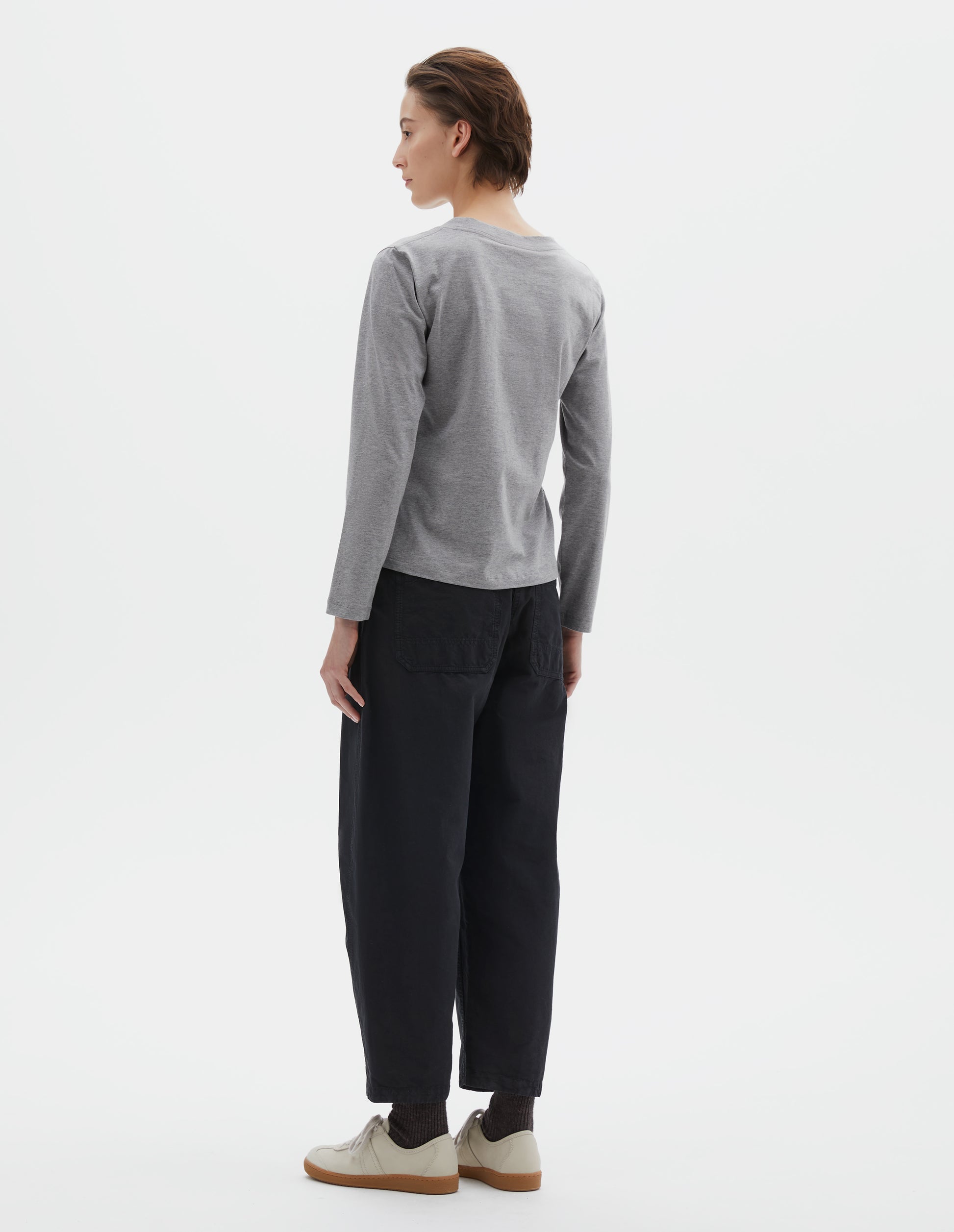 Full body image, back view of model wearing grey cotton wide neck long sleeve with boxy cut. Styled with black cropped trousers, black socks and beige suede trainers.