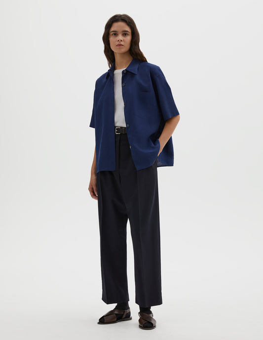 Full body image, front view of model wearing dark blue linen short sleeve shirt, with dropped shoulder, fly front, chest pocket and side split hem. Styled over white cotton t-shirt, with black leather belt, navy blue pleated trousers, black socks and brown leather sandals.