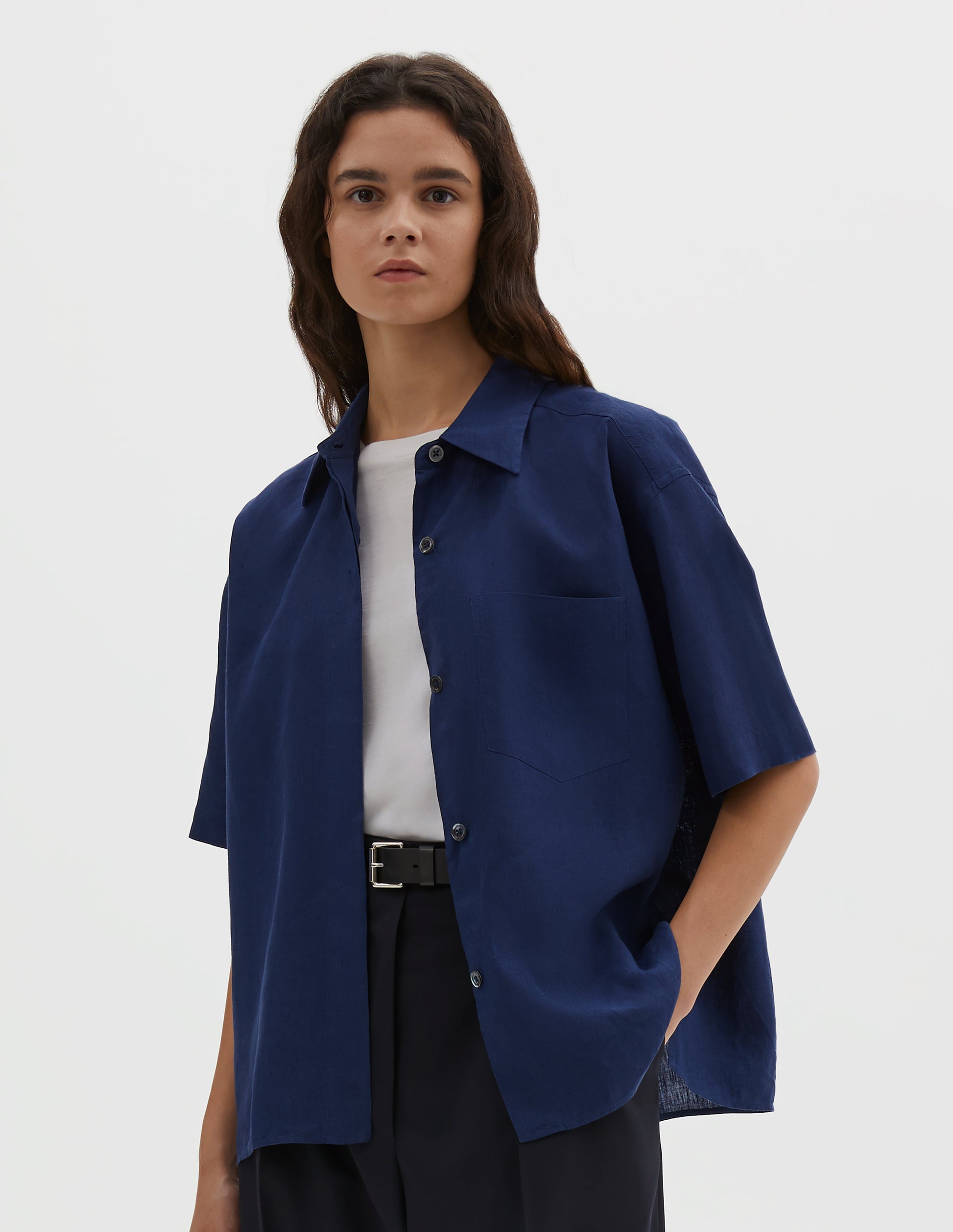 Cropped body image, front view of model wearing dark blue linen short sleeve shirt, with dropped shoulder, fly front, chest pocket and side split hem. Styled over white cotton t-shirt, with black leather belt and navy blue pleated trousers.