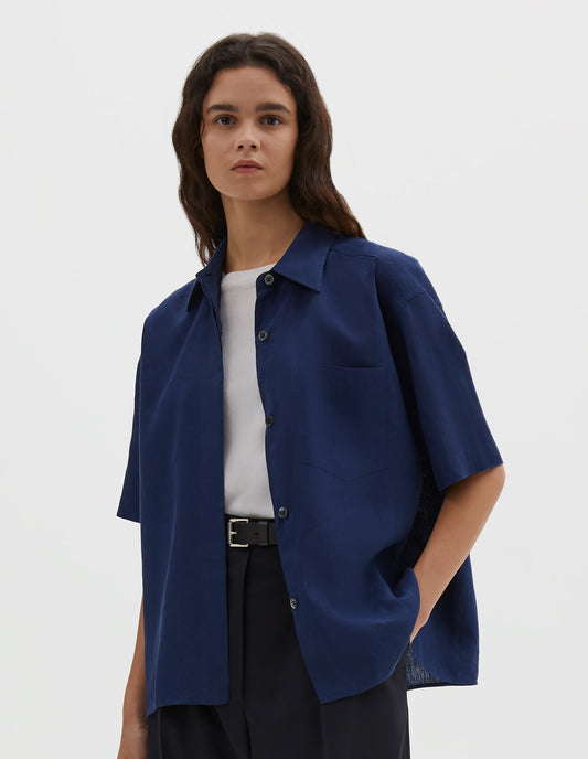 Cropped body image, front view of model wearing dark blue linen short sleeve shirt, with dropped shoulder, fly front, chest pocket and side split hem. Styled over white cotton t-shirt, with black leather belt and navy blue pleated trousers.