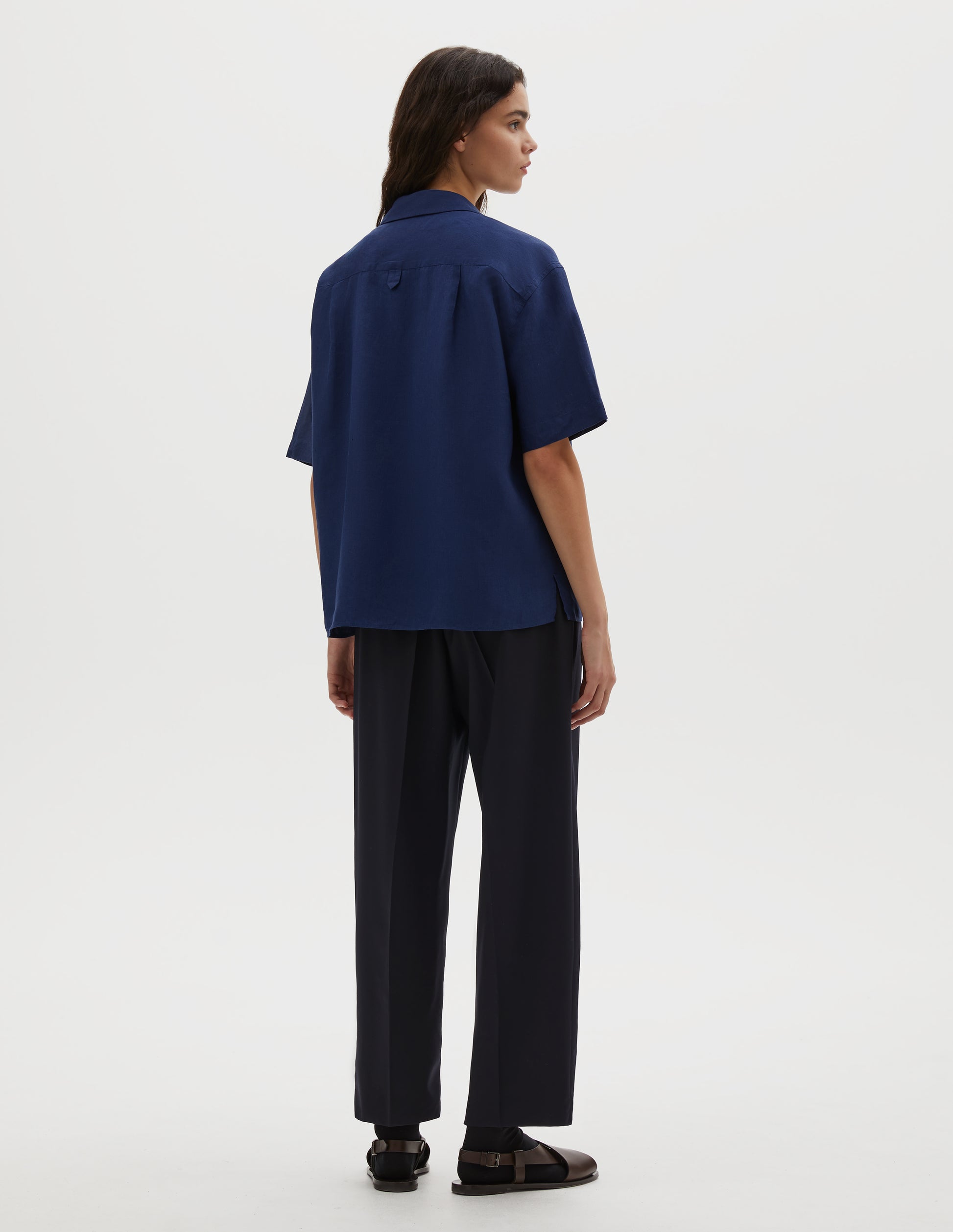 Full body image, back view of model wearing dark blue linen short sleeve shirt, with dropped shoulder, side split hem and hanger loop on back yoke. Styled over white cotton t-shirt, with black leather belt, navy blue pleated trousers, black socks and brown leather sandals.