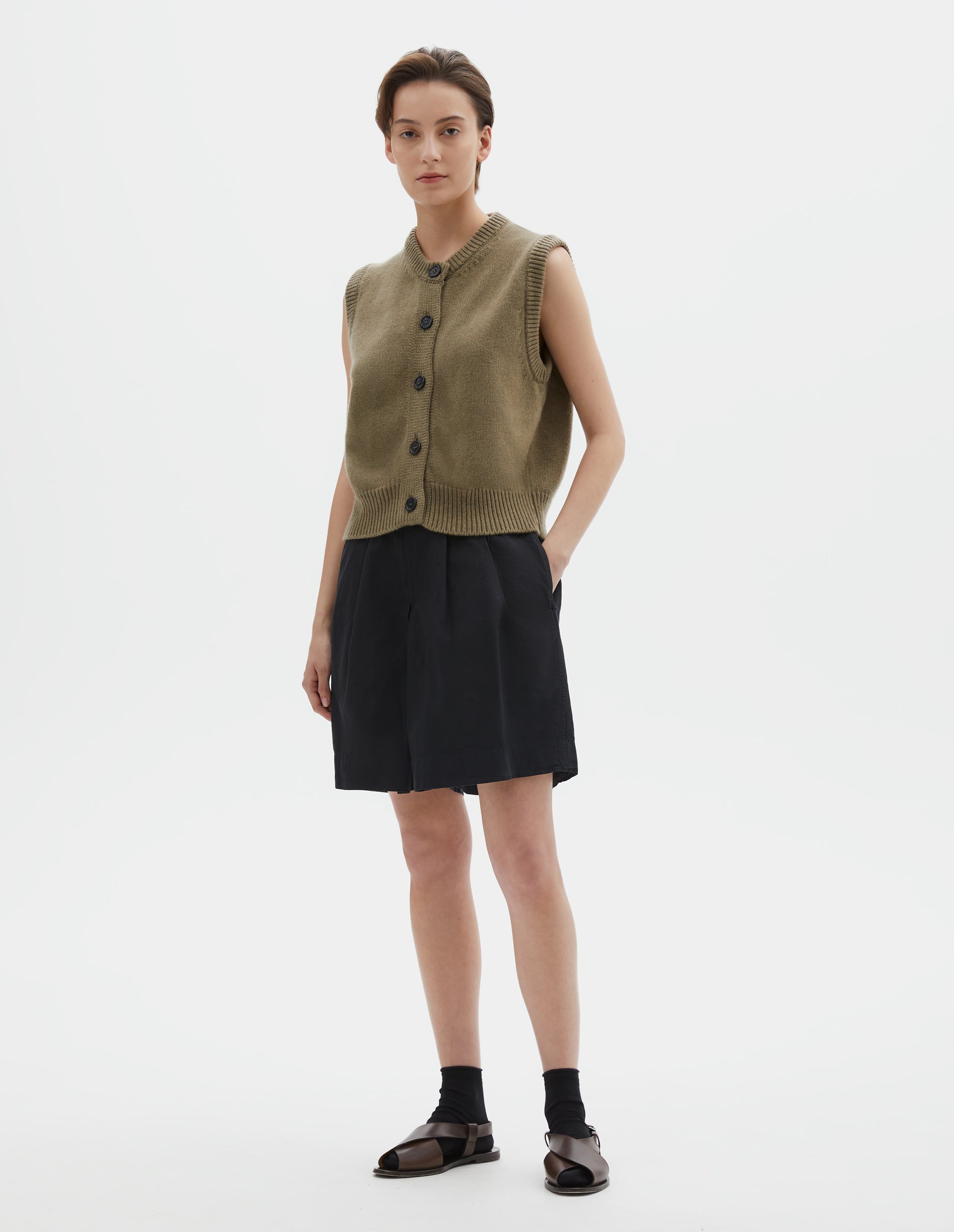 Full body image, front view of model wearing brownish green sleeveless merino cashmere gilet with contrast rib neckline, hem and dark horn buttons. Styled with black shorts, black socks and dark brown leather sandals.