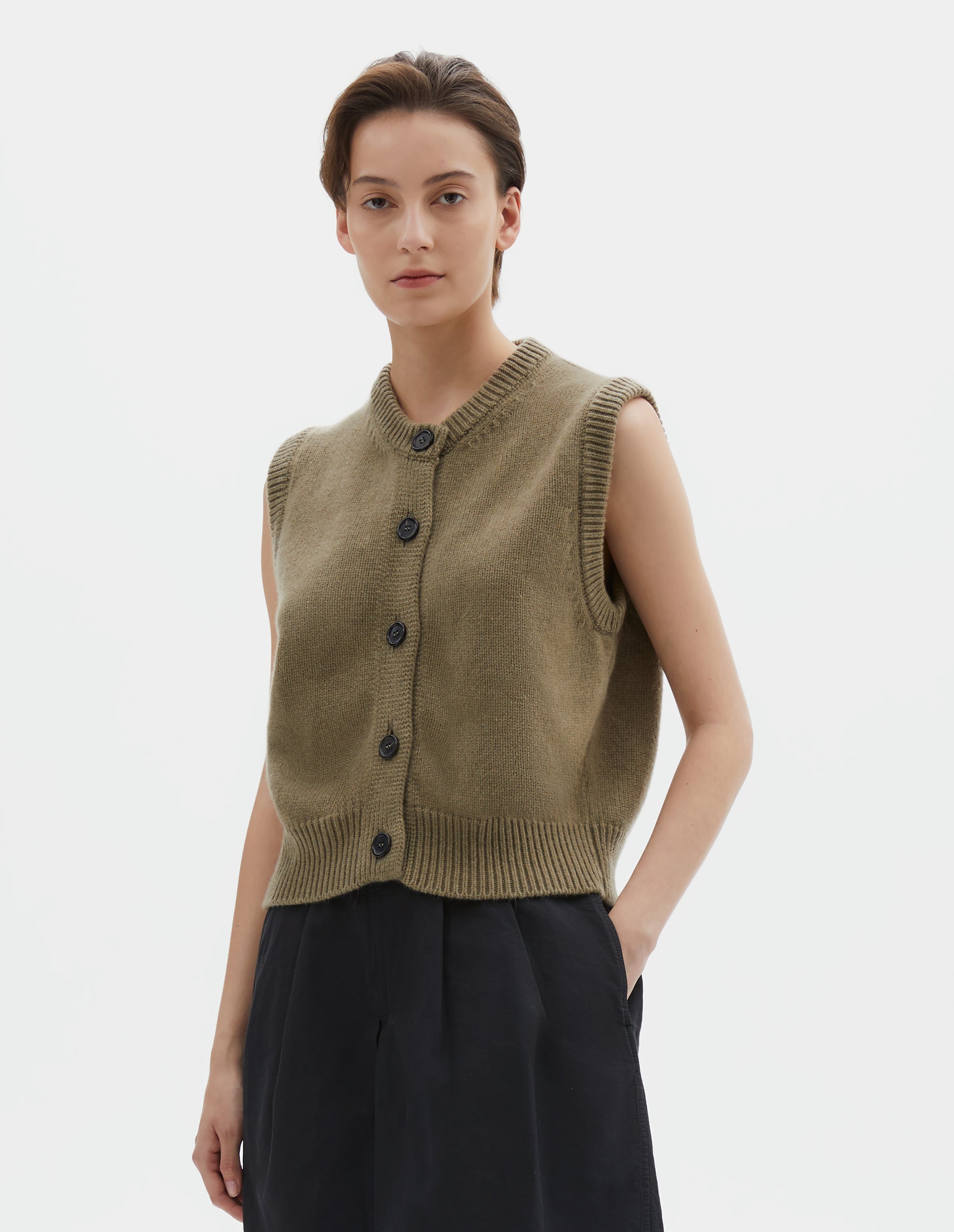 Cropped body image, front view of model wearing brownish green sleeveless merino cashmere gilet with contrast rib neckline, hem and dark horn buttons. Styled with black shorts.