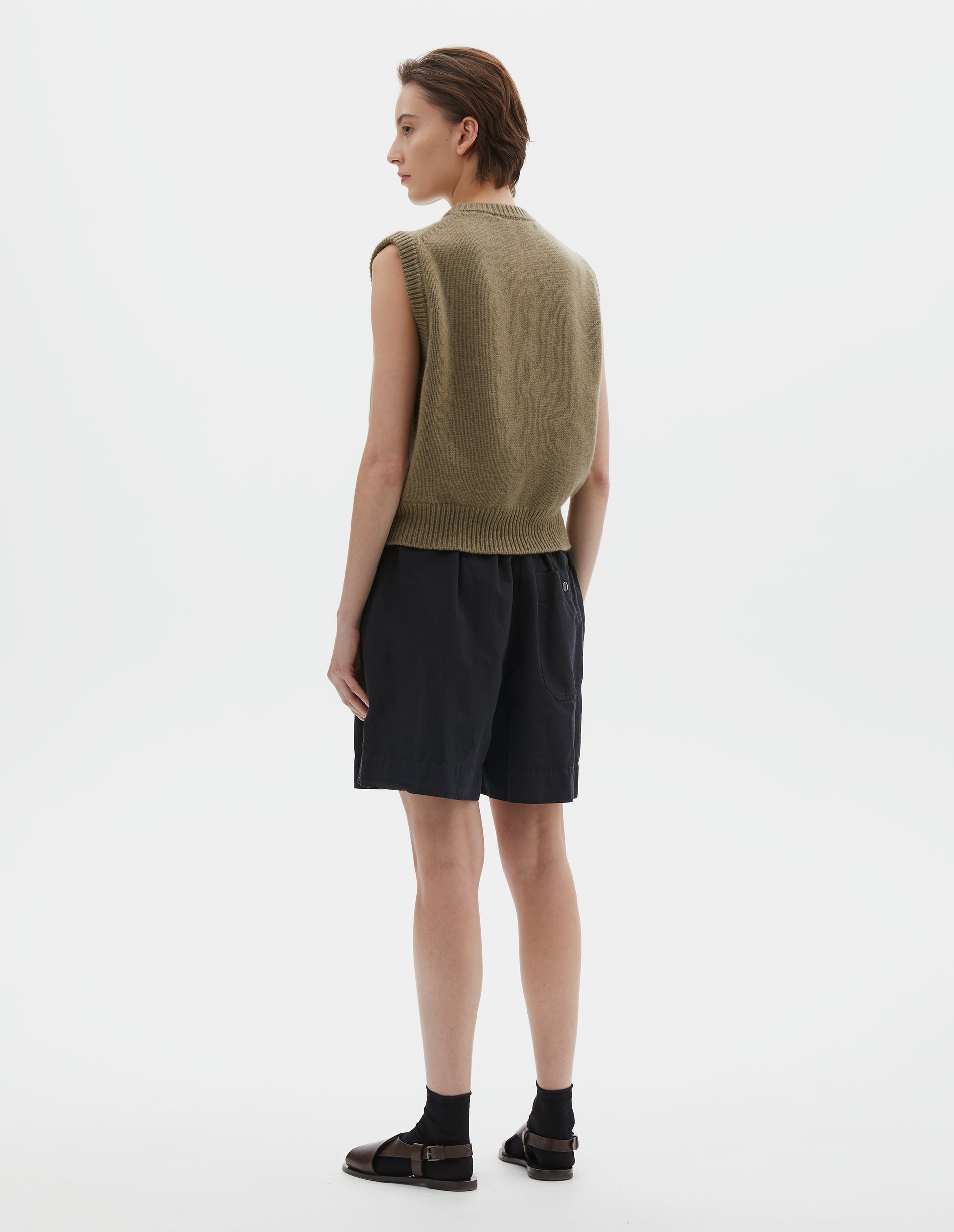 Full body image, back view of model wearing  brownish green sleeveless merino cashmere gilet with contrast rib neckline and hem. Styled with black shorts, black socks and dark brown leather sandals.