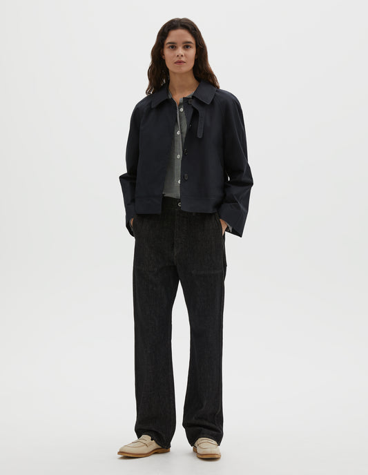 Full body image, front view of model wearing black ventile cotton poplin long sleeve cropped trench, with horn button closure, slant side pockets and adjustable storm tab. Styled over black and white gingham shirt, black denim jeans and with beige loafers.