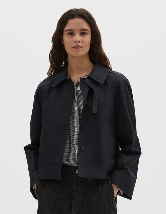 Cropped body image, front view of model wearing black ventile cotton poplin long sleeve cropped trench, with horn button closure, slant side pockets and adjustable storm tab. Styled over black and white gingham shirt and black denim jeans.