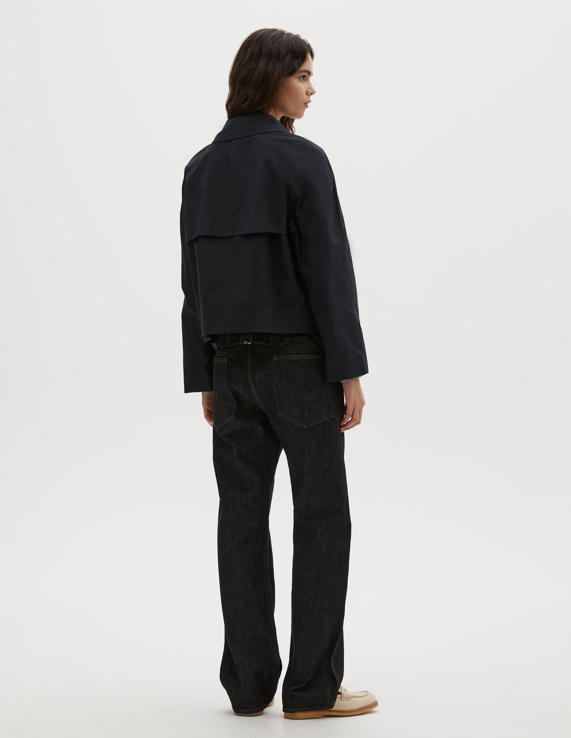 Full body image, back view of model wearing black ventile cotton poplin long sleeve cropped trench, with storm vent. Styled with black denim jeans and beige loafers.