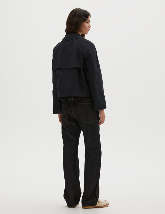 Full body image, back view of model wearing black ventile cotton poplin long sleeve cropped trench, with storm vent. Styled with black denim jeans and beige loafers.