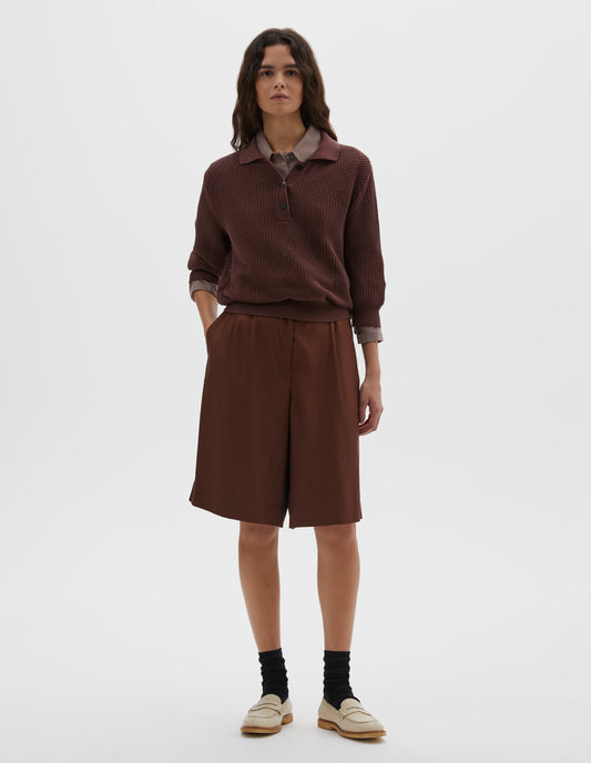 Full body image, front view of model wearing brownish red cotton cashmere pointelle polo shirt, with solid seam detailing, rib collar, placket, cuffs and hem and horn button closure. Styled over pink round collar shirt, with dark red shorts, black socks and beige suede loafers.