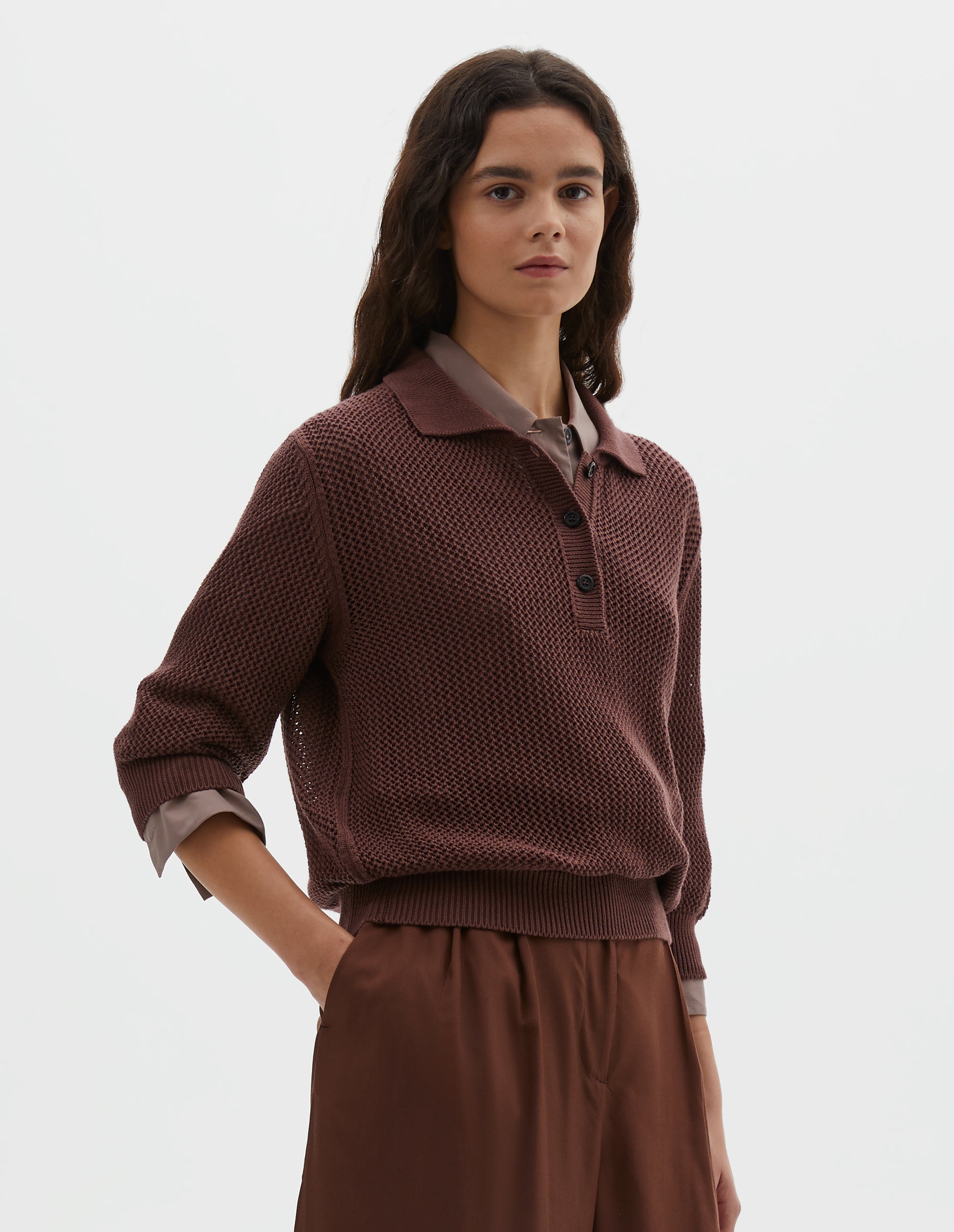 Cropped body image, front view of model wearing brownish red cotton cashmere pointelle polo shirt, with solid seam detailing, rib collar, placket, cuffs and hem and horn button closure. Styled over pink round collar shirt, with dark red shorts.
