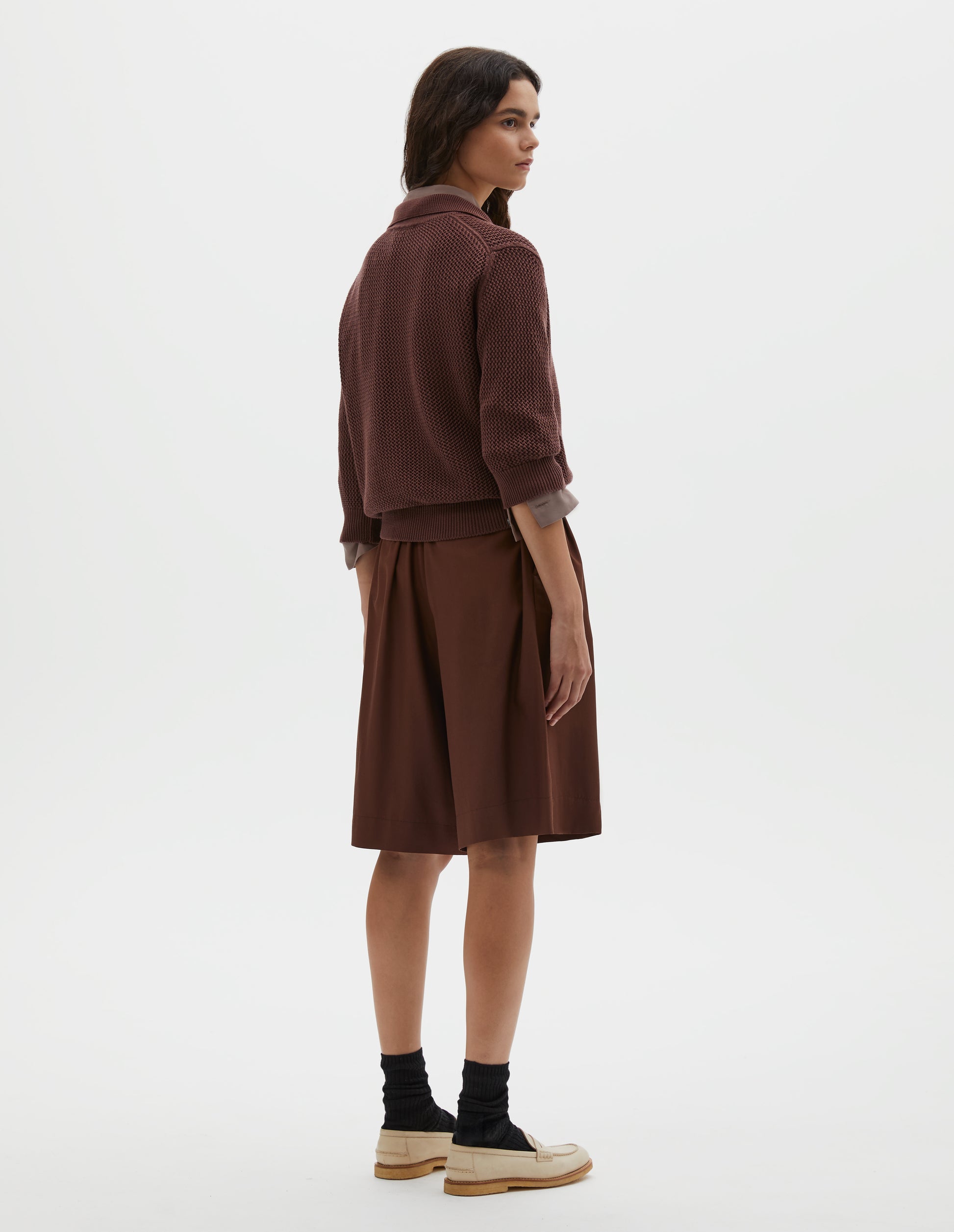 Full body image, back view of model wearing brown regular cut knee length shorts with with half-elasticated waist, slant side pockets and welt back pocket. Styled with dark pink shirt, brown knitted polo, black socks and beige leather loafers.