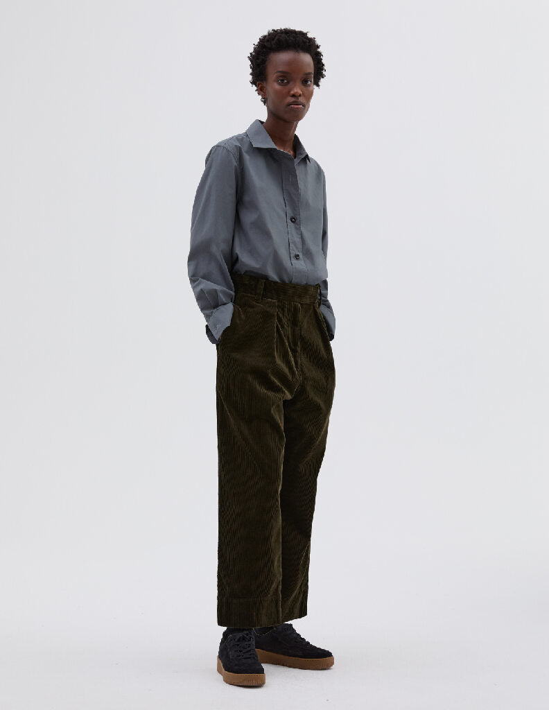 Full body image, front view of model wearing dark green corduroy cropped trousers, with zip fly with hook and bar tab fastening, single pleats, belt loops, concealed side pockets and deep stitch hem. Styled with blue shirt tucked in and black suede trainers. 