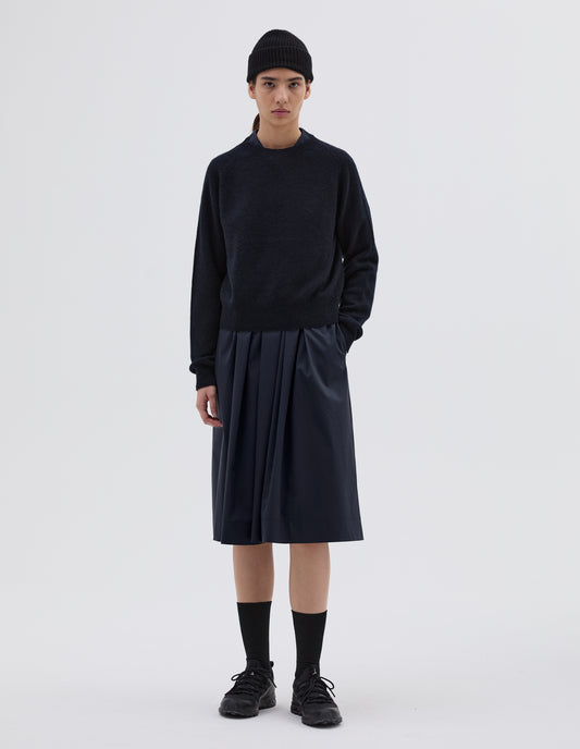 Full body image, front view of model wearing dark navy jumper with raglan sleeve, contrast rib on neck, cuffs and hem. Styled with navy blue beanie, pleated navy blue skirt, black socks and black trainers.