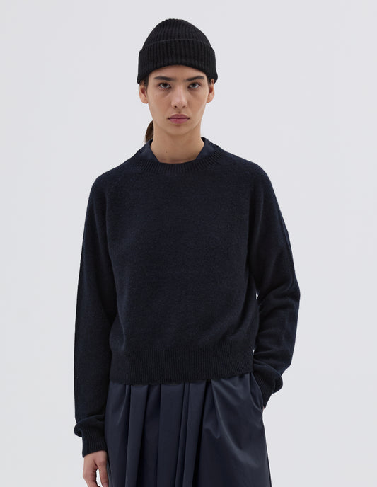 Cropped body image, front view of model wearing dark navy jumper with raglan sleeve, contrast rib on neck, cuffs and hem. Styled with navy blue beanie and pleated navy blue skirt.