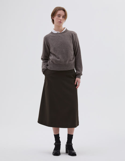Full body image, front view of model wearing greyish beige jumper with raglan sleeve, contrast rib on neck, cuffs and hem. Styled over white shirt, with brown skirt, black socks and black open toe heels.