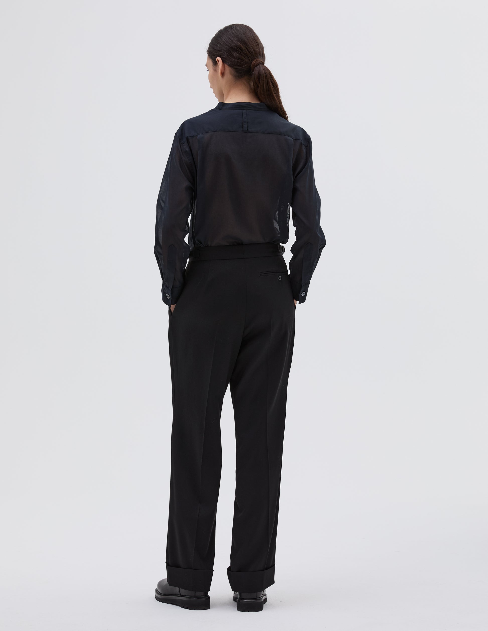 Full body image, back view of model wearing black wool wide leg trousers, with button through jet back pocket. Styled with navy blue collarless shirt tucked in and black leather boots.