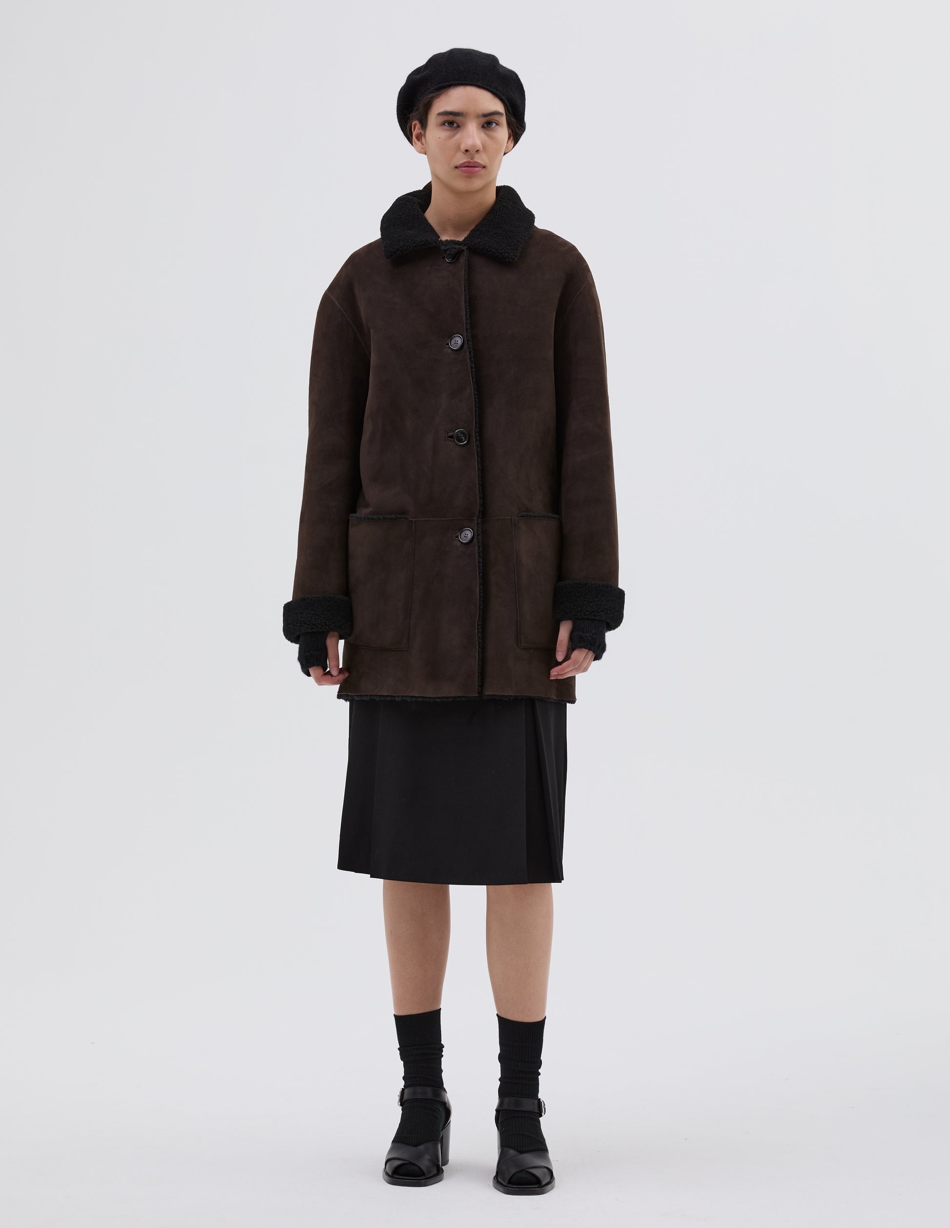 Full body image, front view of model wearing brown and black merino sheepskin car coat with storm tab on collar, patch pockets and horn button closure. Styled with black hat, skirt, socks and black leather heels.