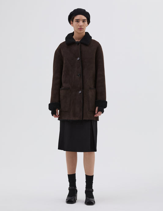 Full body image, front view of model wearing brown and black merino sheepskin car coat with storm tab on collar, patch pockets and horn button closure. Styled with black hat, skirt, socks and black leather heels.