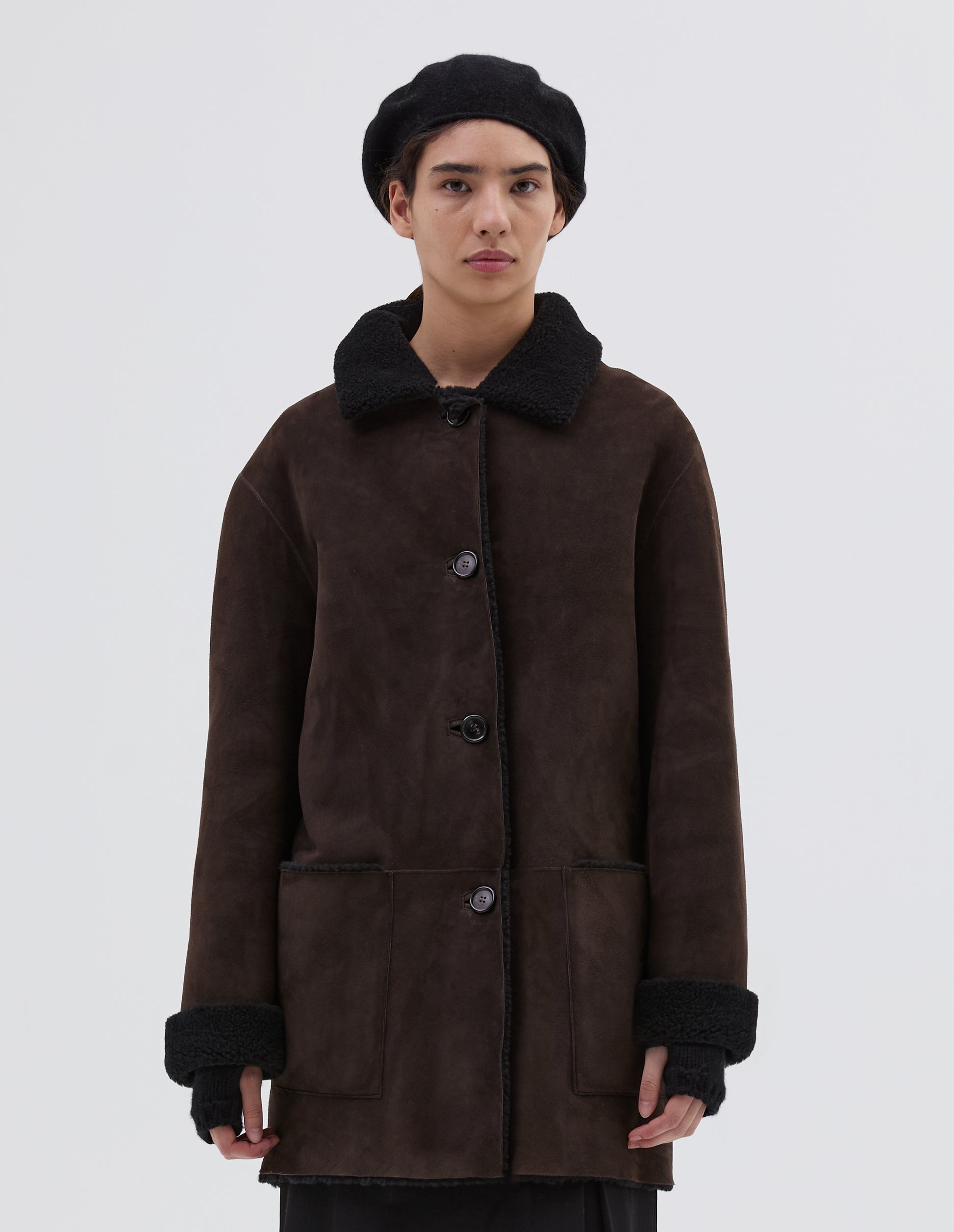 Cropped body image, front view of model wearing brown and black merino sheepskin car coat with storm tab on collar, patch pockets and horn button closure. Styled with black hat and skirt.