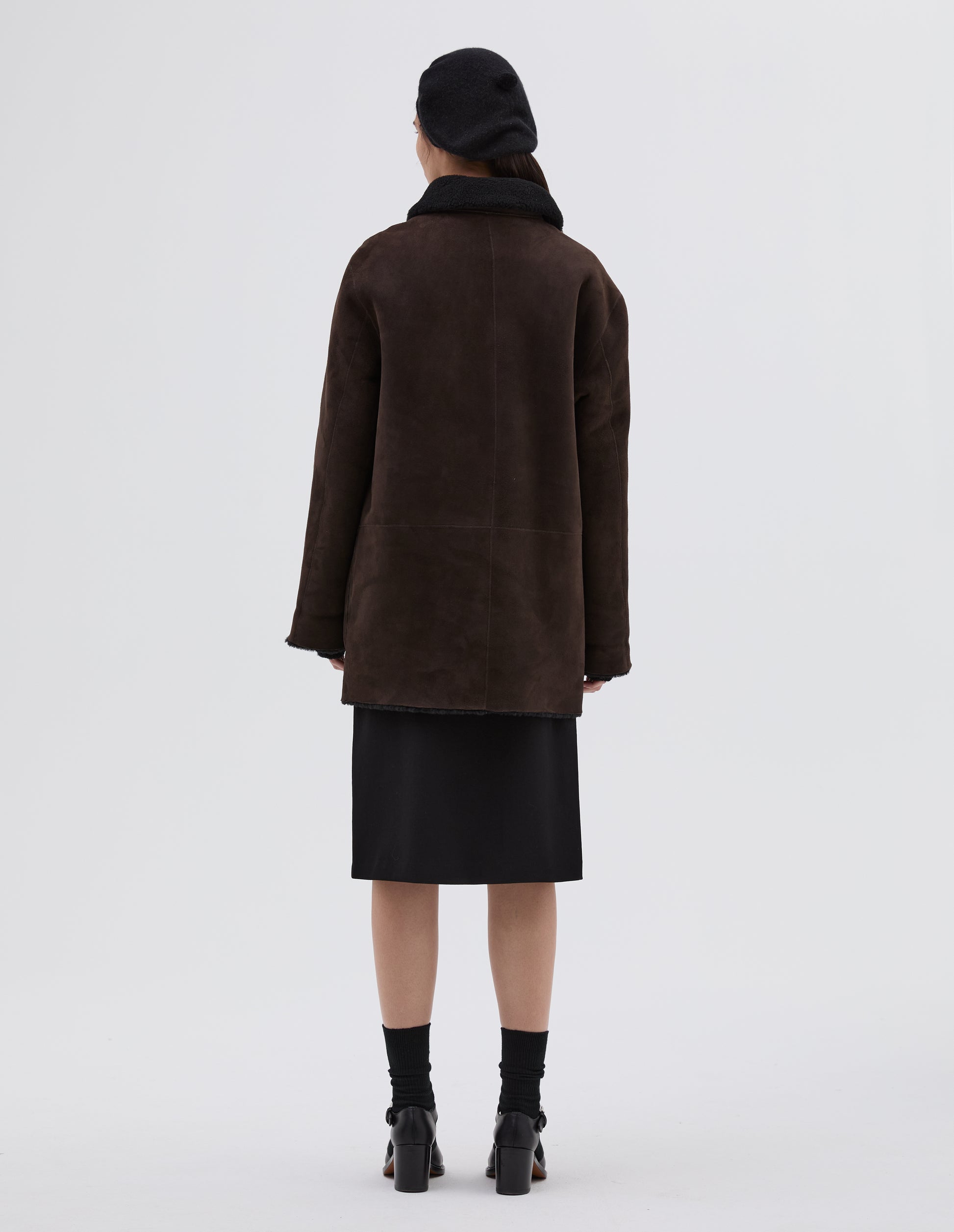 Full body image, back view of model wearing brown and black merino sheepskin car coat contrast collar and cuffs. Styled with black hat, skirt, socks and black leather heels.