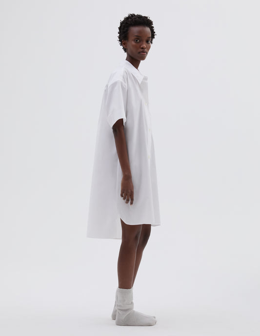 Full body image, side view of model wearing white lightweight cotton short sleeve nightshirt with mother of pearl buttons, patch pocket and side split hem with longer back. Styled with light grey bed socks.