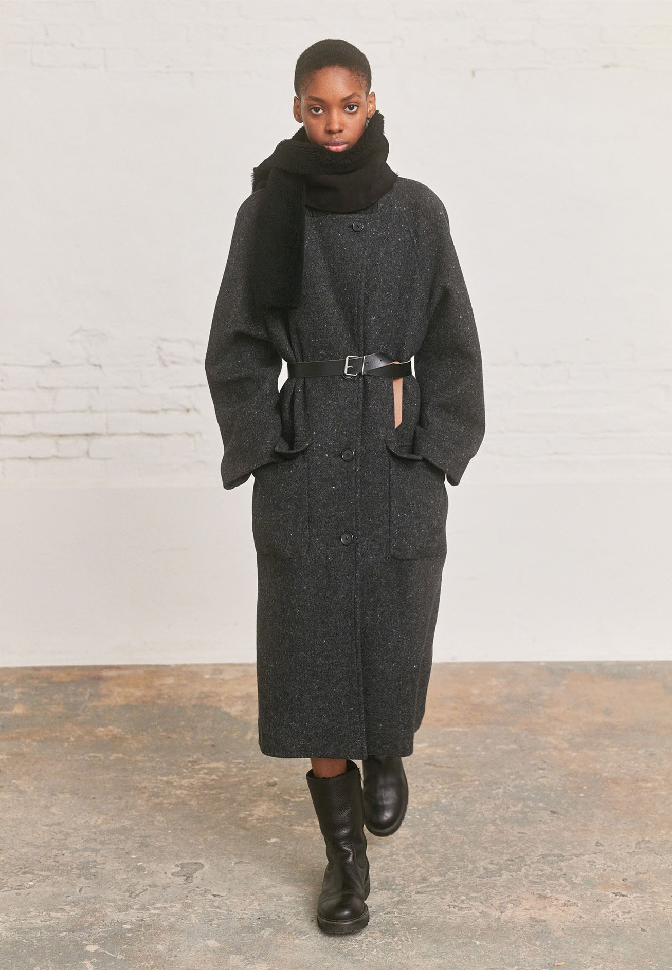 Full body image, front view of model wearing black tie trough shearling scarf. Styled with dark grey wool coat with leather belt and black leather boots.