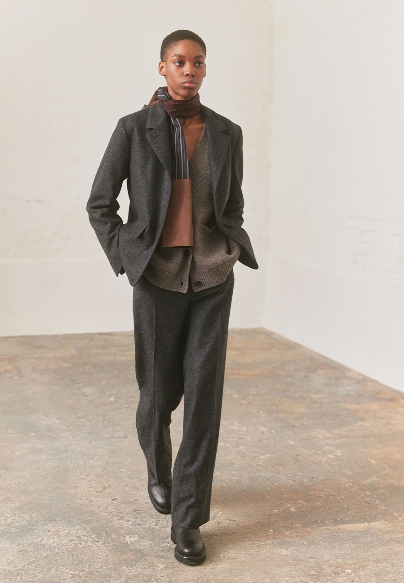 Full body image, front view of model wearing dark grey blazer with horn buttons on lapel and cuffs and flap patch pockets. Styled over beige knitted cardigan, with matching trousers, red and blue shirting scarf and black leather boots.