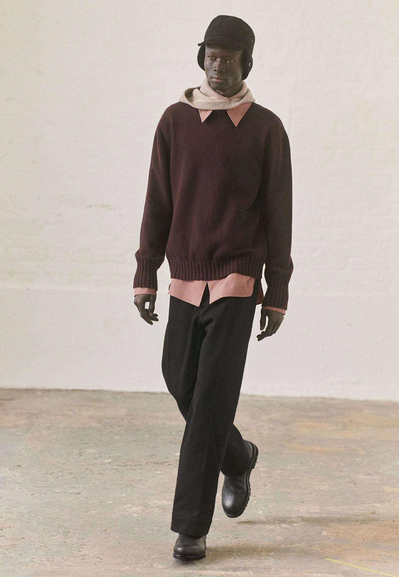 Full body image, front view of model wearing black press stud shearling cap with earflaps down. Styled with beige neckwarmer and pink shirt under brown jumper, black trousers and black leather boots.