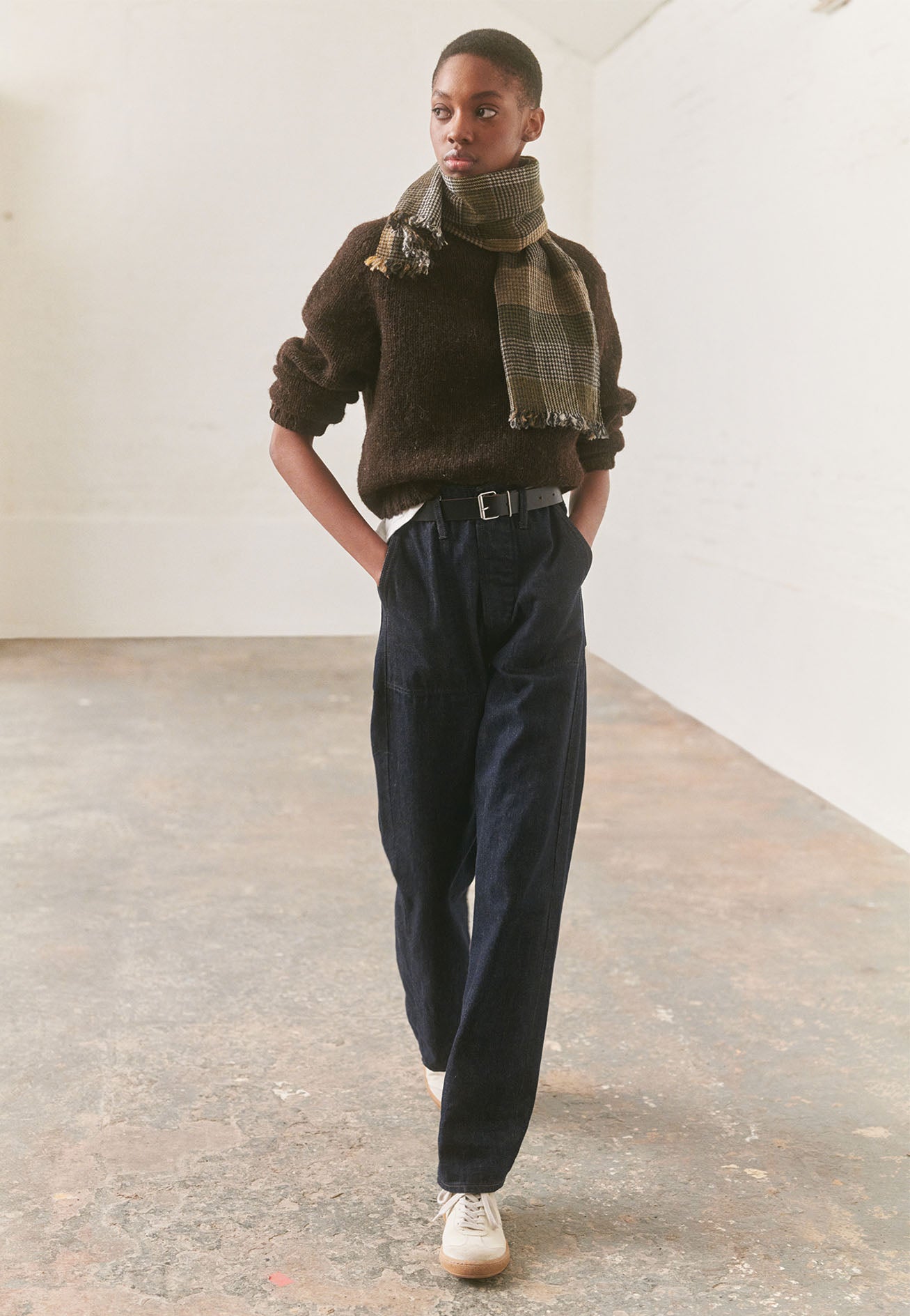 Full body image, front view of model wearing dark brown rustic wool jumper with saddle sleeve and contrasting rib on neck, cuffs and hem. Styled with green, beige and black checked scarf, black leather belt, indigo blue jeans and white trainers with gum sole.