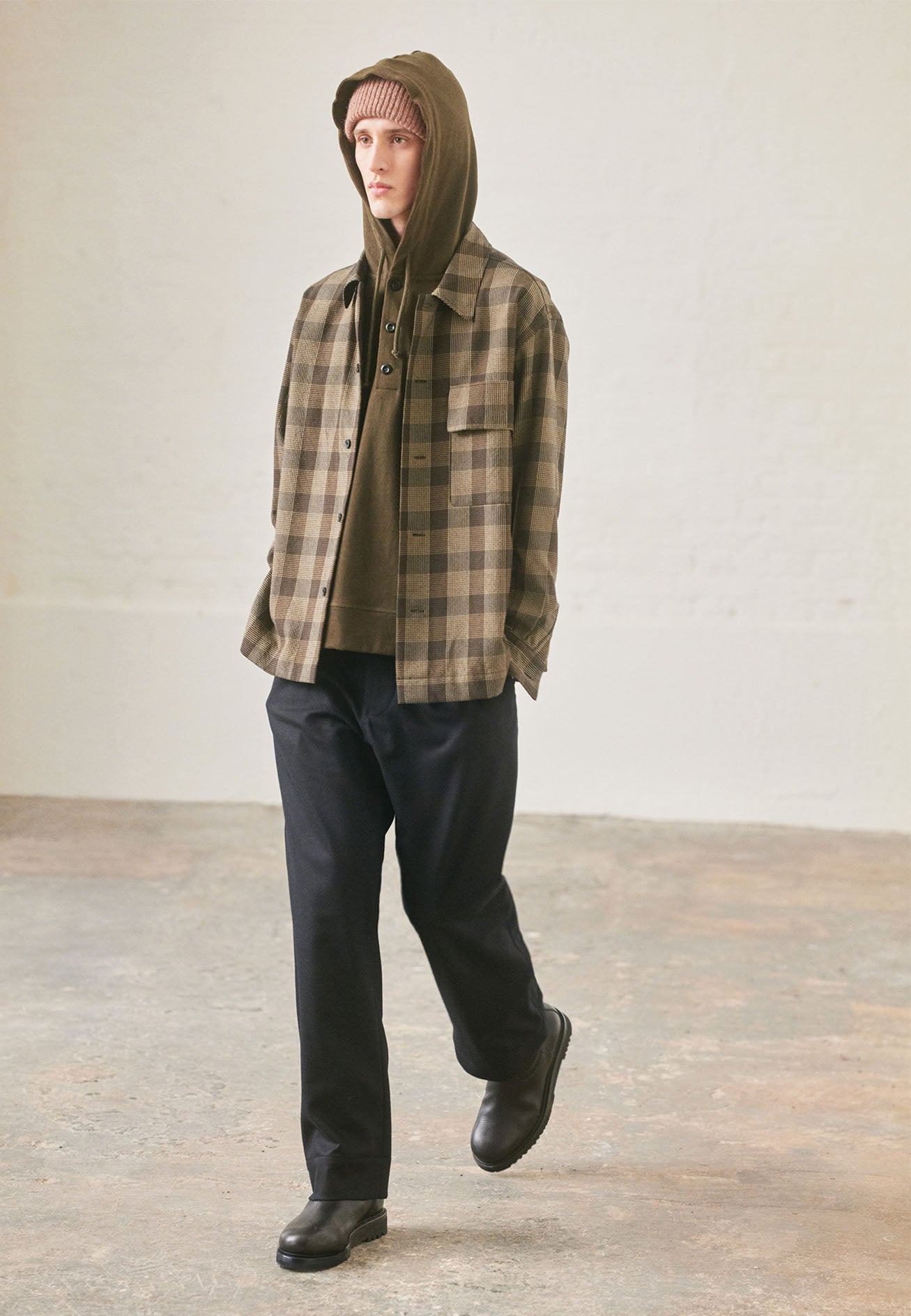 Full body image, front view of model wearing black, brown and green checked wool shirt with horn button fastening, deep placket and flap patch pocket. Styled with khaki green button hoodie, dark navy trousers and black leather boots.