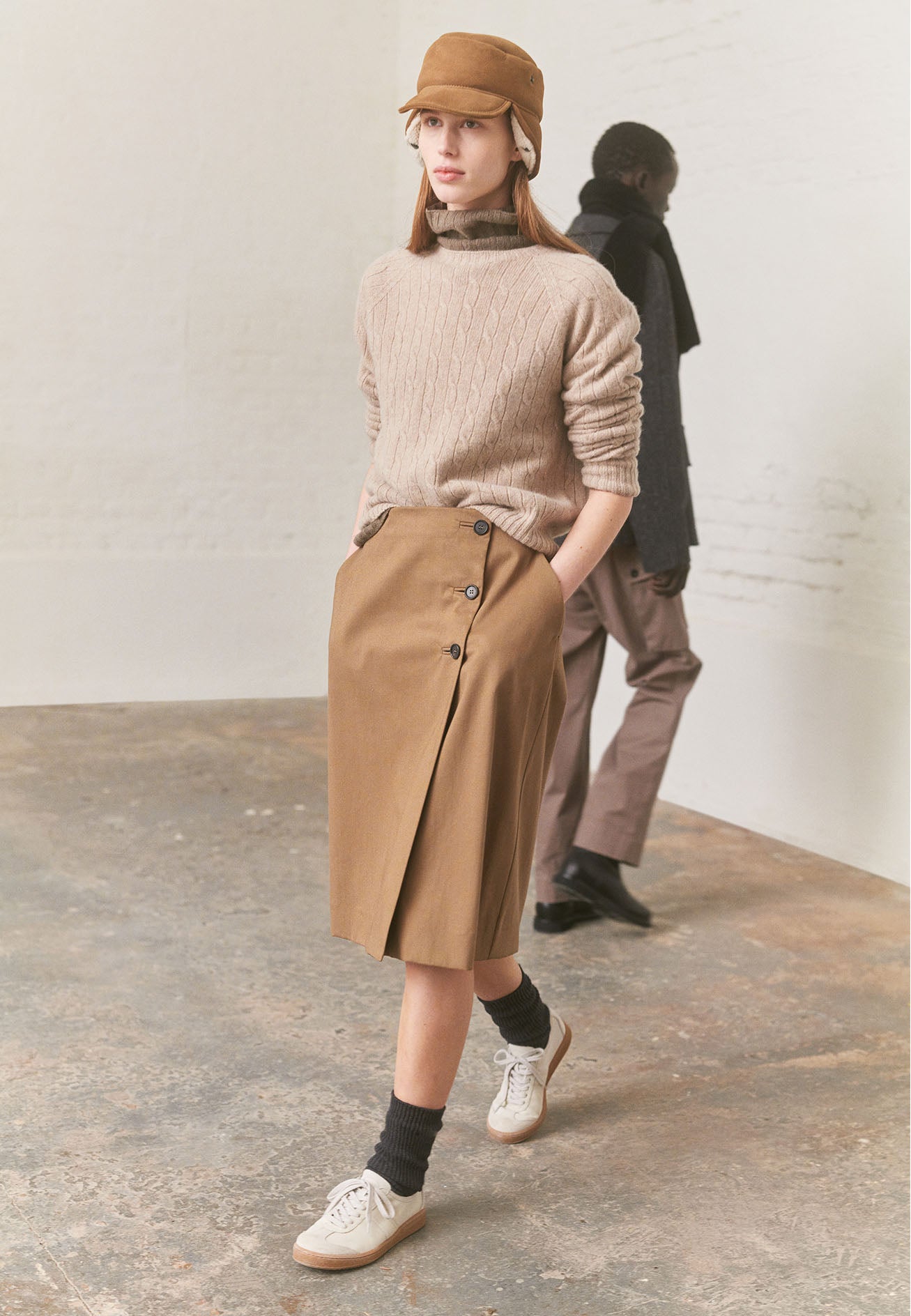 Full body image, front view of model wearing a beige knee length skirt, with horn button fastening, deep pleat and concealed side pockets. Styled with beige cable knit jumper, beige shearling hat, grey socks and white trainers with gum sole.
