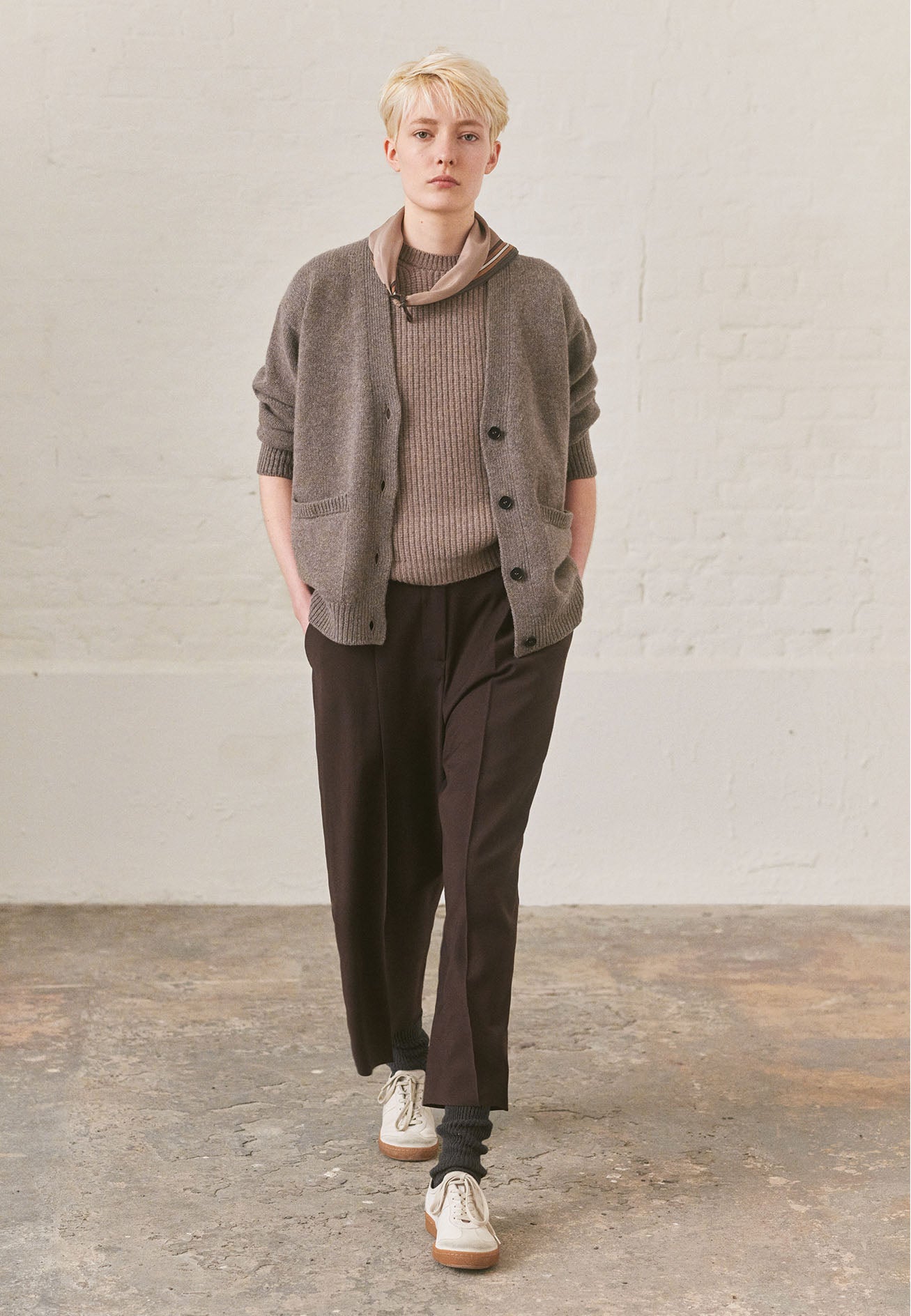 Full body image, front view of model wearing beige v-neck cardigan, with contrast rib on placket, cuffs and hem and two front pockets, finished with horn button closure. Styled over beige knitted top, with beige silk scarf, brown trousers, grey socks and white trainers with gum sole.