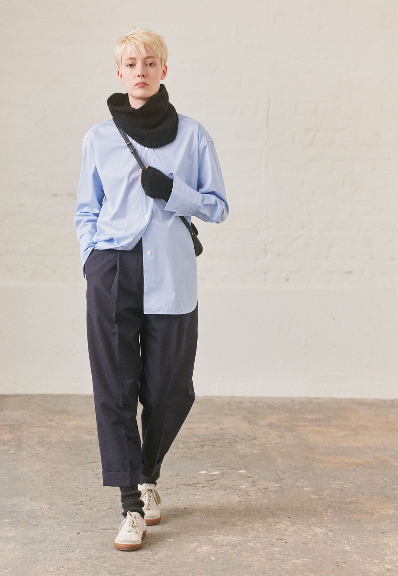 Full body image, front view of model wearing relaxed light blue shirt with wide raised placket and mother of pearl buttons, and turned up cuffs. Styled with navy pinstripe trousers, black knitted neck warmer, black fingerless gloves, black leather bag and white trainers with gum sole.