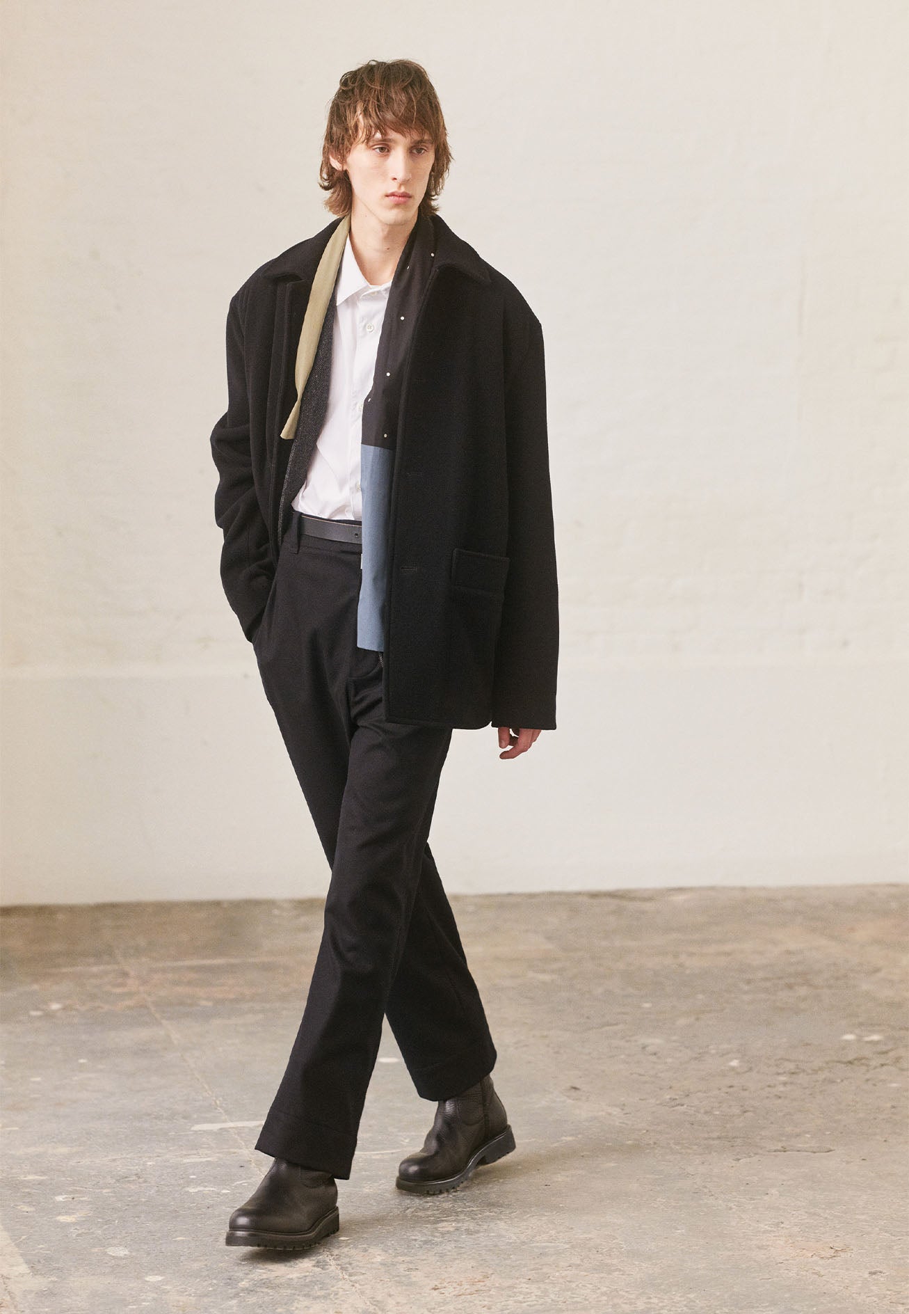 Full body image, front view of model wearing navy blue trousers, with half elasticated waistband, belt loops, horn button tab fastening and slant pockets. Styled with white shirt, blue and beige shirting scarf, black car coat and black leather boots.