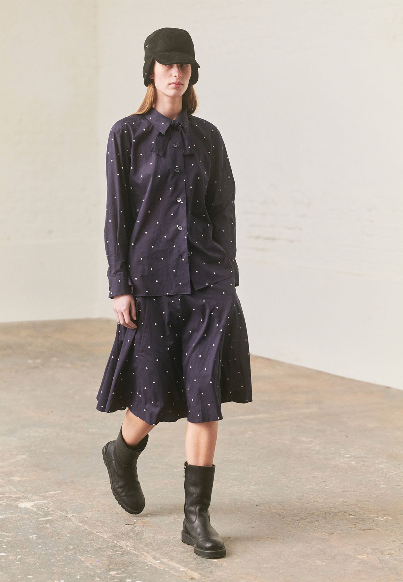 Full body image, front view of model wearing navy and white polka dot long sleeve shirt with wide stitch through placket, mother of pearl buttons, elongated back and removeable self fabric tie. Styled with matching skirt, black shearling hat and black leather boots.