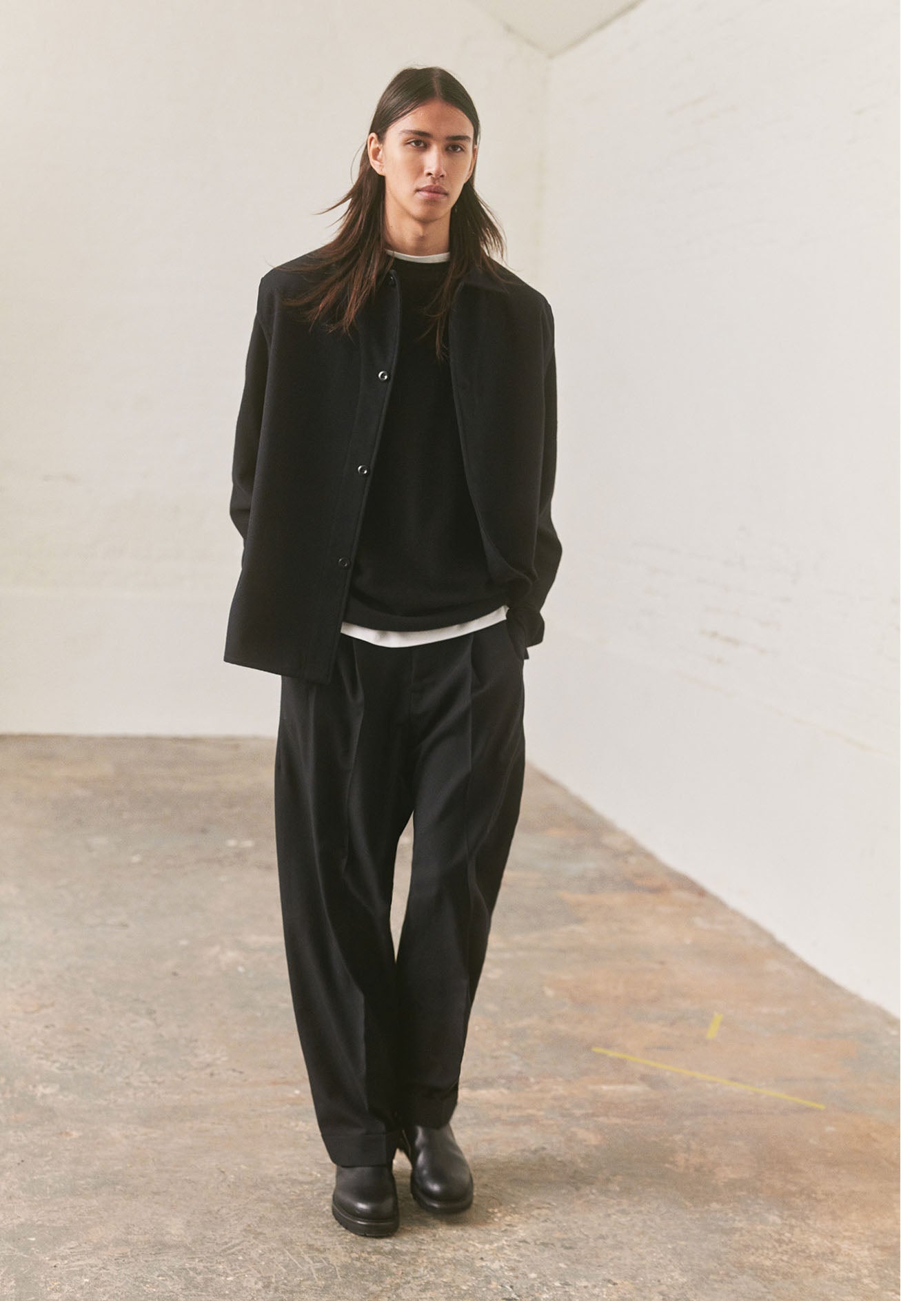 Full body image, front view of model wearing relaxed black crew neck jumper with rolled edge detailing on neck, rib trim on cuffs and hem. Styled underneath a black wool shirt with black pleated trousers and black leather boots.
