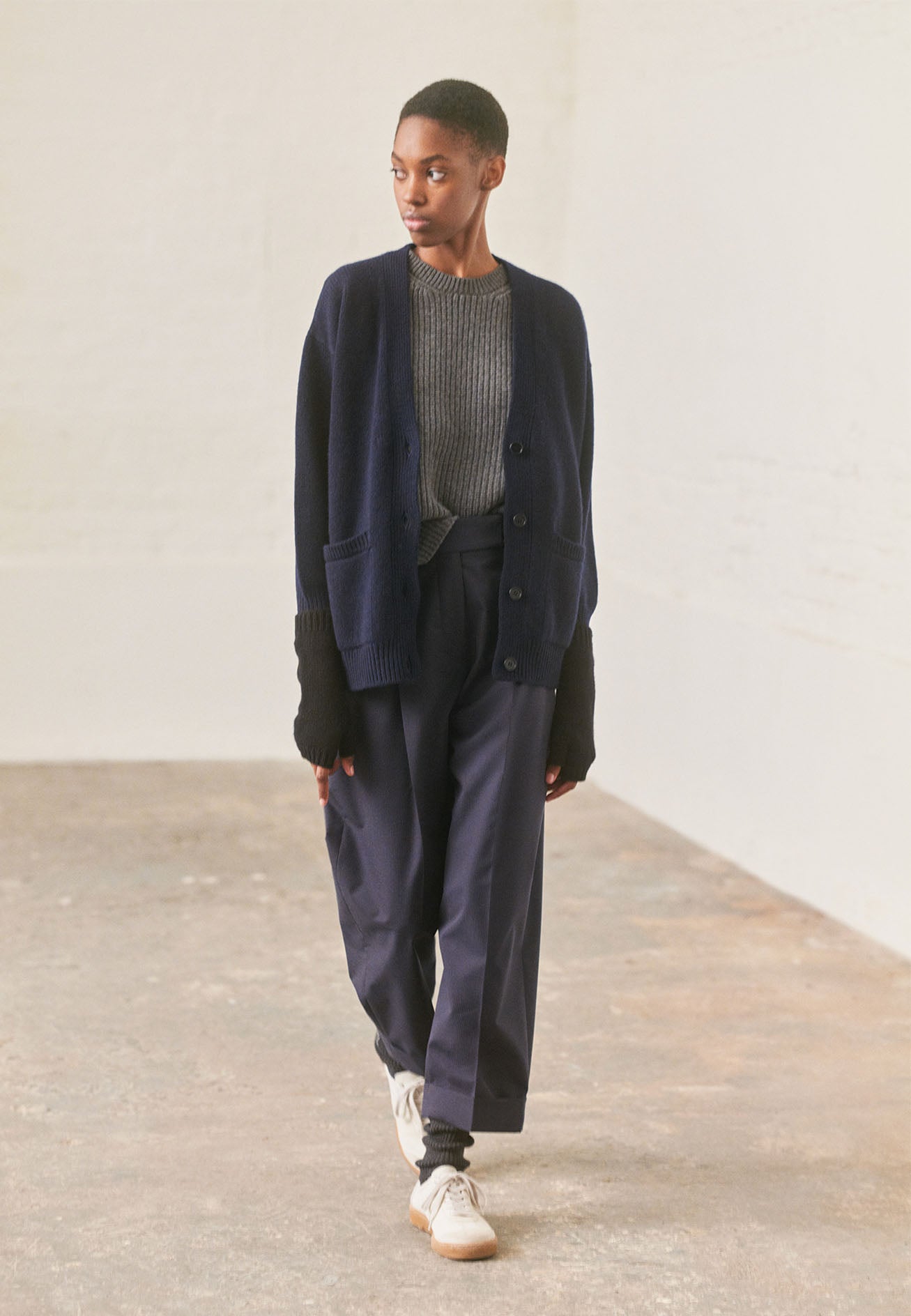 Full body image, front view of model wearing navy v-neck cardigan, with contrast rib on placket, cuffs and hem and two front pockets, finished with horn button closure. Styled over grey knitted top, with navy blue pinstripe trousers, grey socks and white trainers with gum sole.