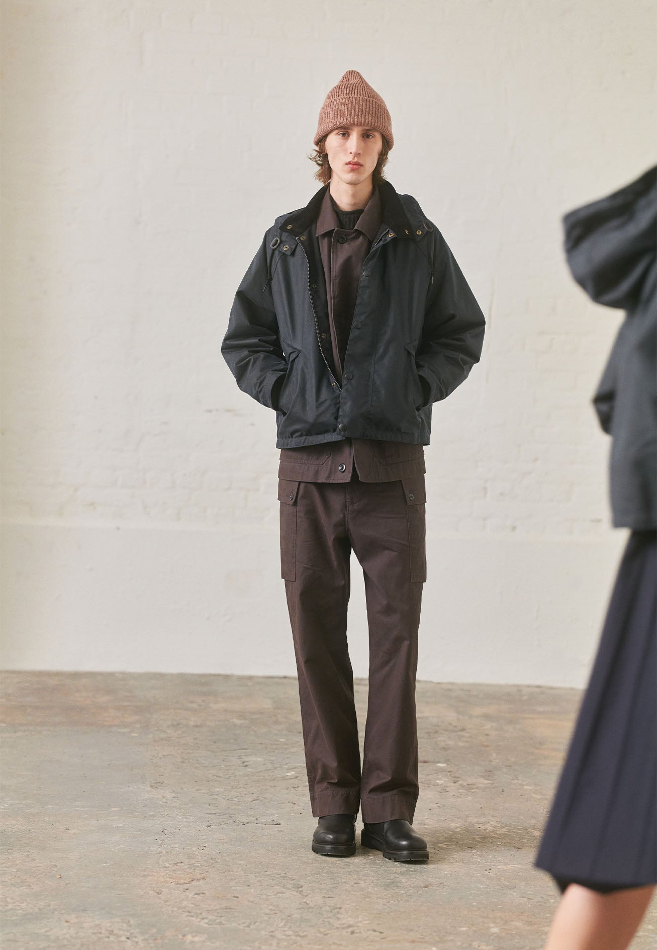 Full body image, front view of model wearing brown raglan sleeve jacket with corozo button fastening fly front. Styled underneath navy cropped coat, with matching brown cargo trousers, pale pink knitted hat and black leather boots.