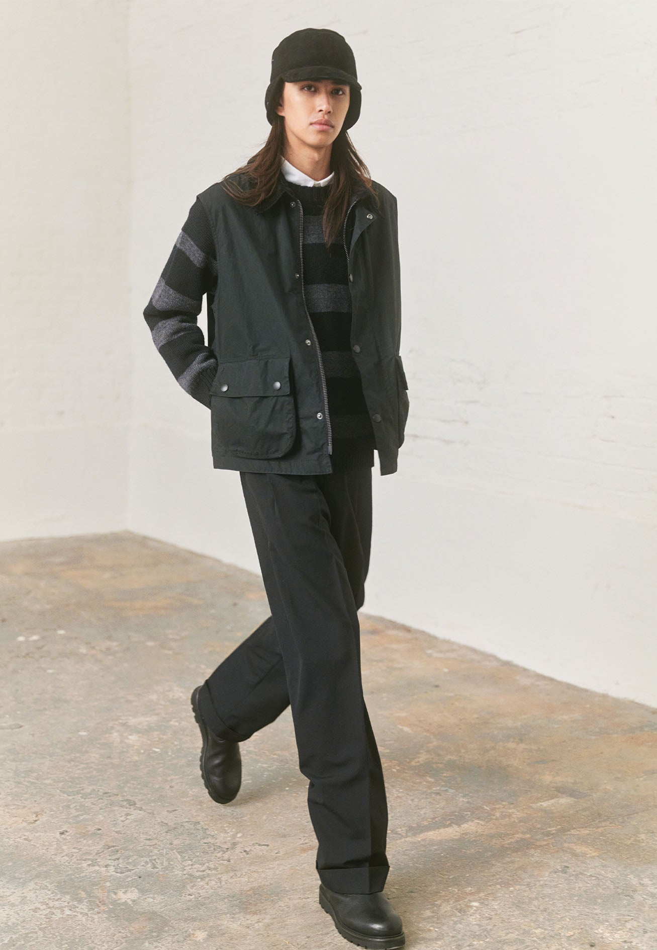Full body image, front view of model wearing black relaxed gilet with corduroy collar, two-way zip fastening and press stud fastening storm flap, and large bellows pockets. Styled with grey and black striped jumper, white shirt, black trousers, black shearling hat and black leather boots.
