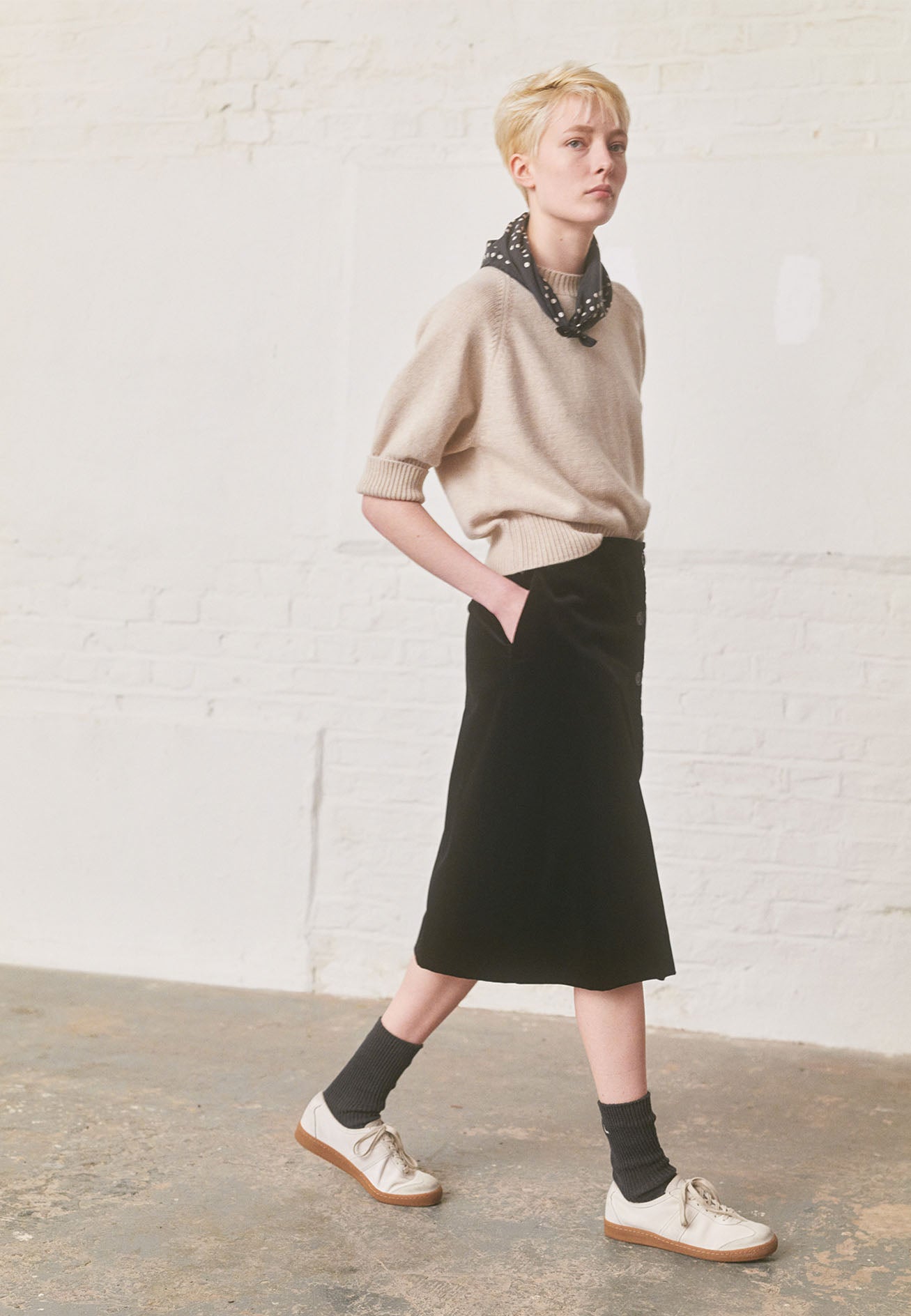 Full body image, front view of model wearing black velvet a-line skirt with deep wrap, horn button fastening and slant side pockets. Styled with beige short sleeve jumper, black and white polka dot scarf, green socks and white trainers with gum sole.