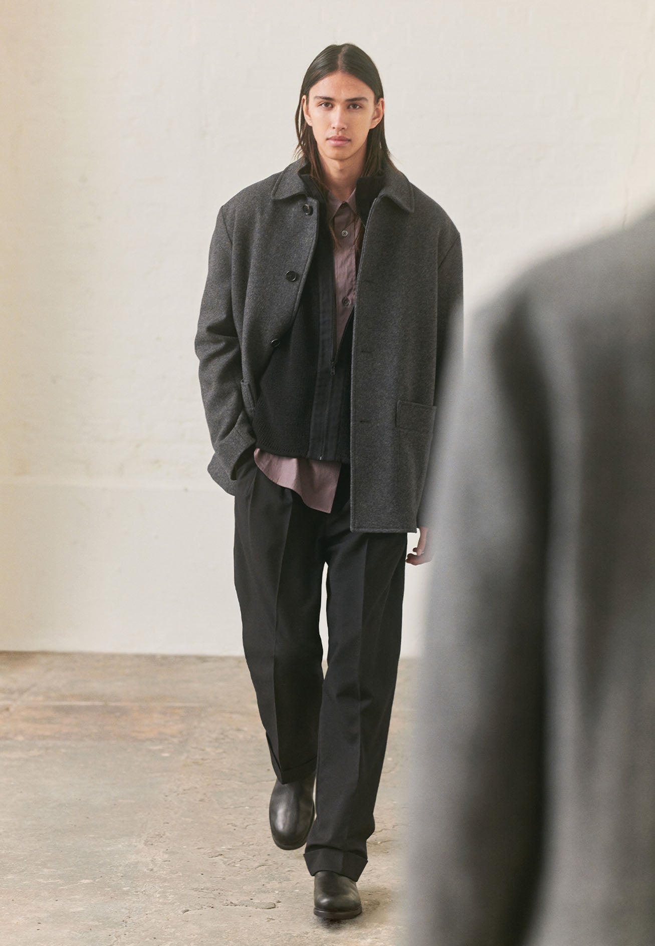 Full body image, front view of model wearing grey shirt with patch pocket and button-through flap pocket on chest, raised placket with mother of pearl buttons. Styled with black cardigan, charcoal grey car coat, black trousers and black leather boots.