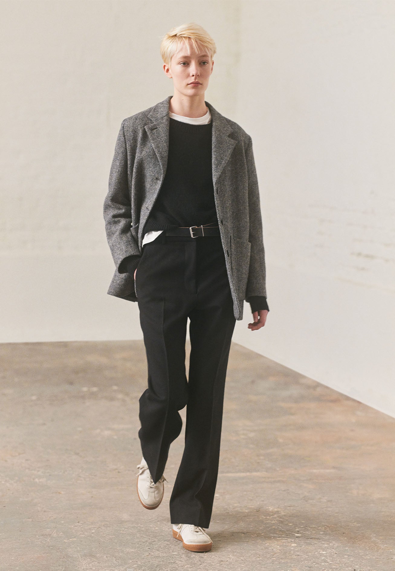 Full body image, front view of model wearing relaxed grey and black herringbone  blazer with horn buttons and large patch pockets. Styled over white long sleeve t-shirt and black jumper. With black leather belt, black trousers and white trainers with gum sole.