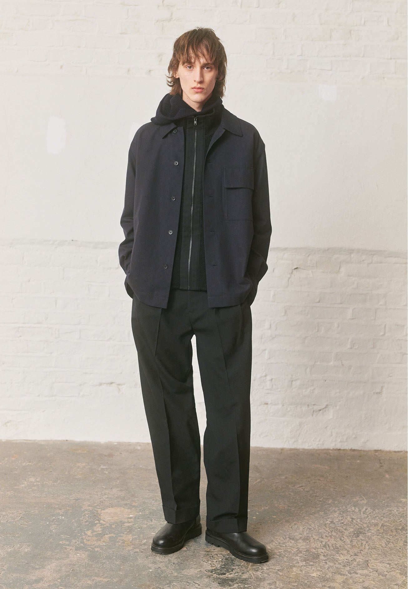 Full body image, front view of model wearing black zip through wool cardigan with stand collar, straight hem and contrast cotton tape placket with chunky Vislon zip fastening. Styled with dark navy overshirt, charcoal grey hood, black trousers and black leather boots.