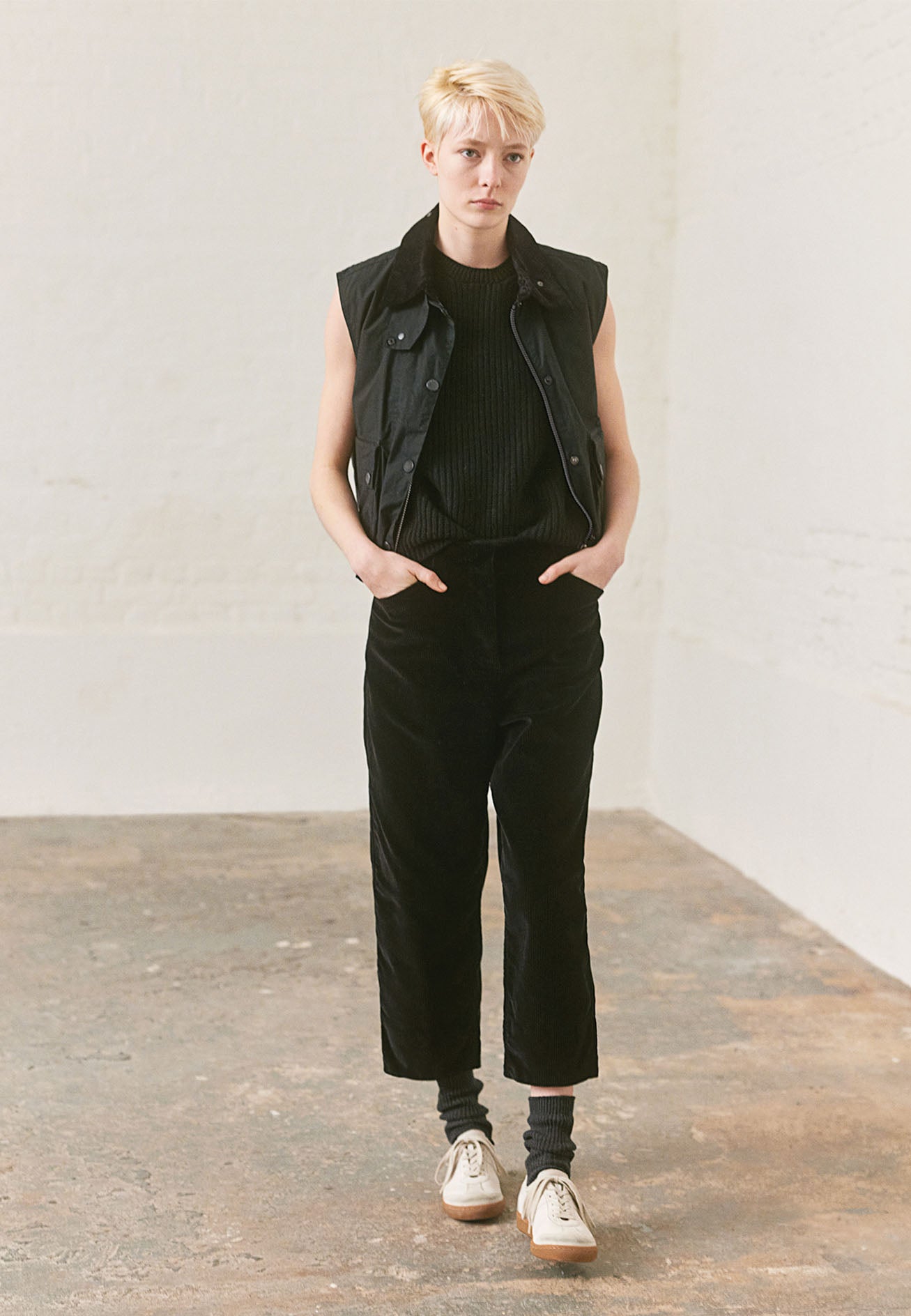 Full body image, front view of model wearing black waxed gilet with contrast corduroy collar, zip fastening, press stud storm flap and bellows envelope patch pockets. Styled with dark navy knitted vest, black velvet trousers, grey socks and white trainers with gum sole. 