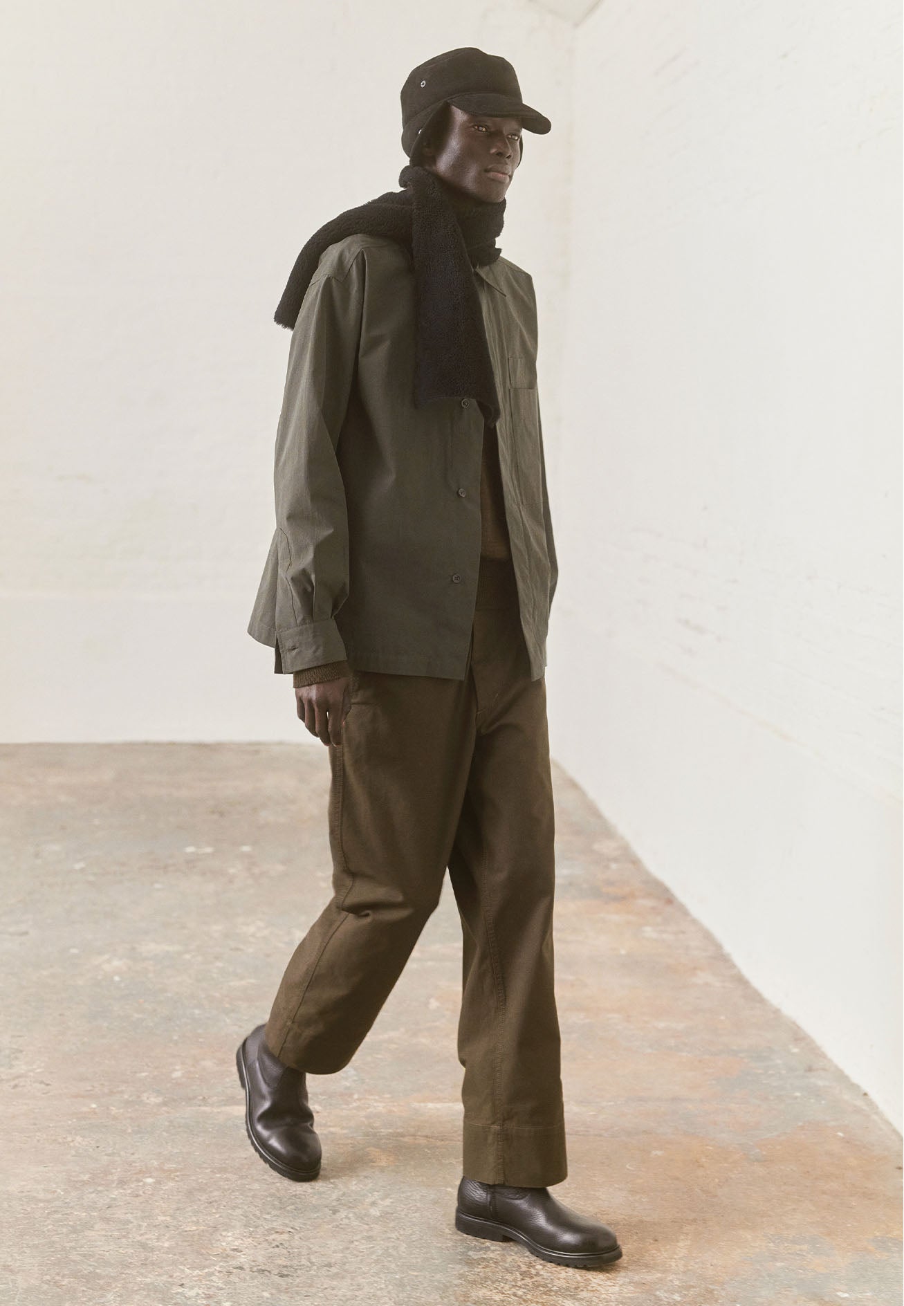 Full body image, front view of model wearing dark green shirt with wide placket, large patch chest pocket with internal pen pocket. Styled with khaki brown trousers, black shearling scarf and hat and black leather boots.