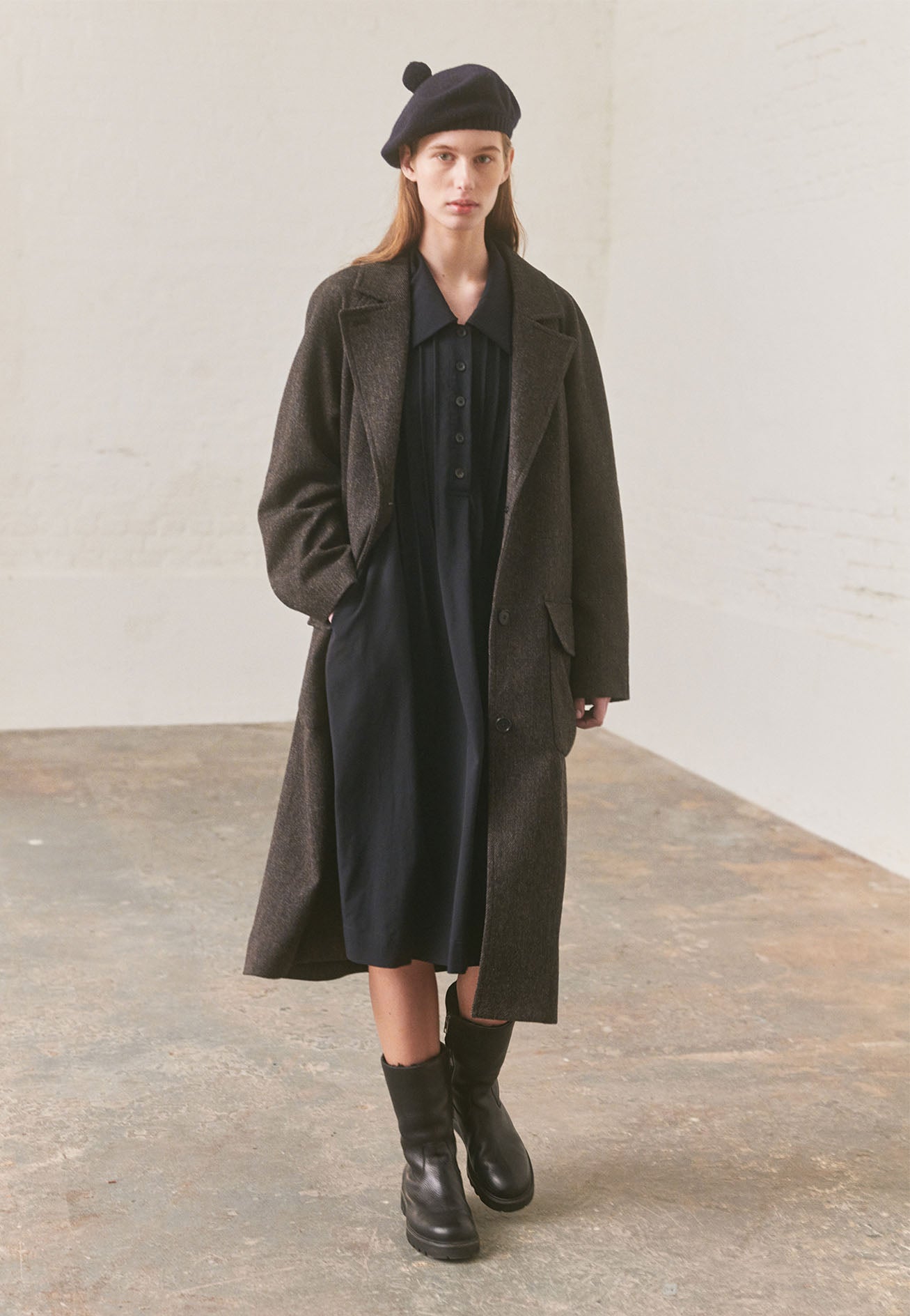Full body image, front view of model wearing dark navy collared dress with pleat detailing and horn button closure. Styled with brown overcoat, navy knitted bobble hat and black leather boots.